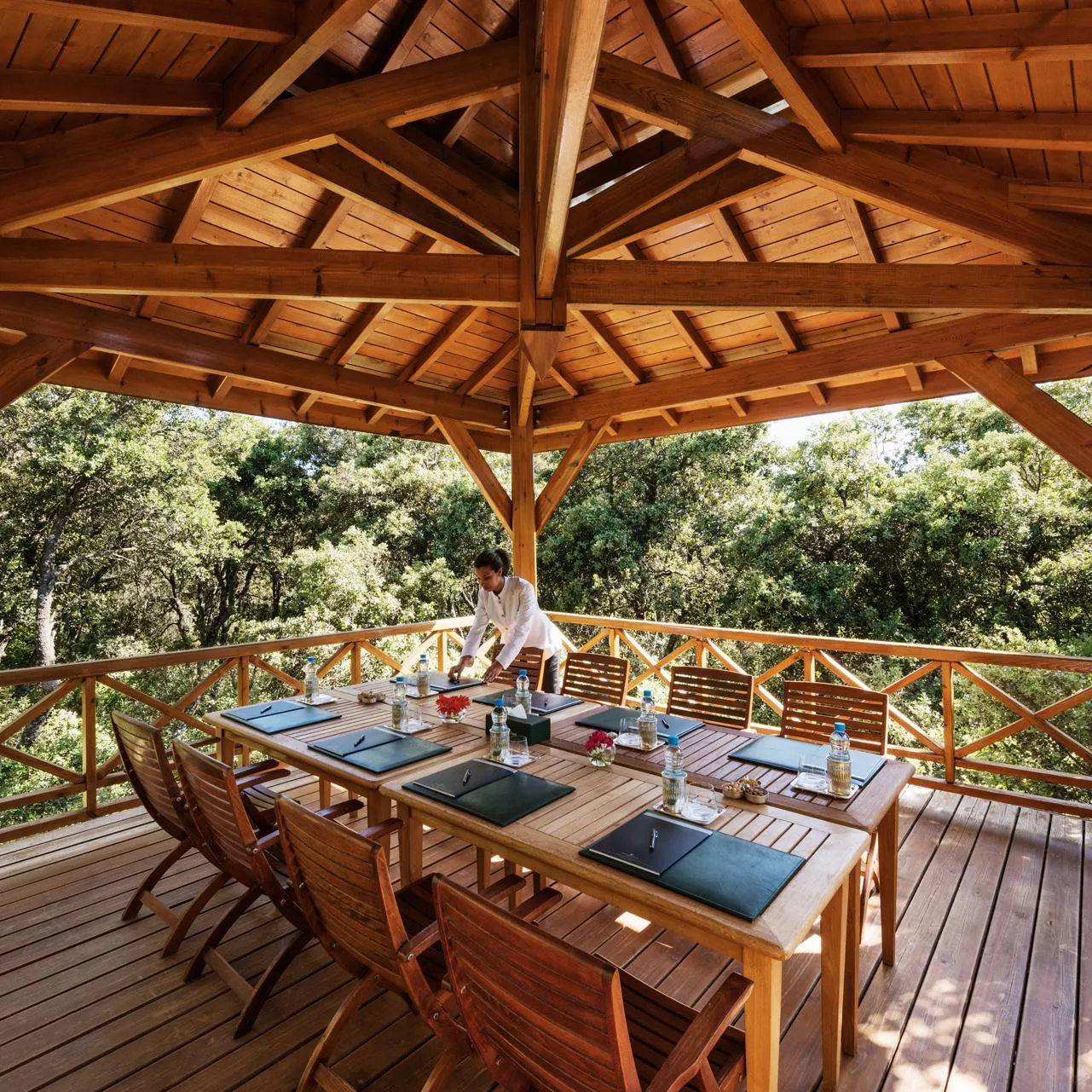 A person prepares tables in an outdoor wooden structure surrounded by trees.
