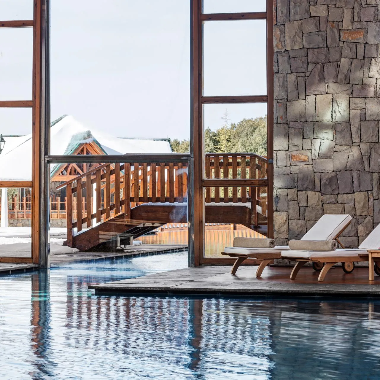 Two deck chairs with towels and drinks are placed near an indoor pool, with a view of a snowy exterior through large glass windows.
