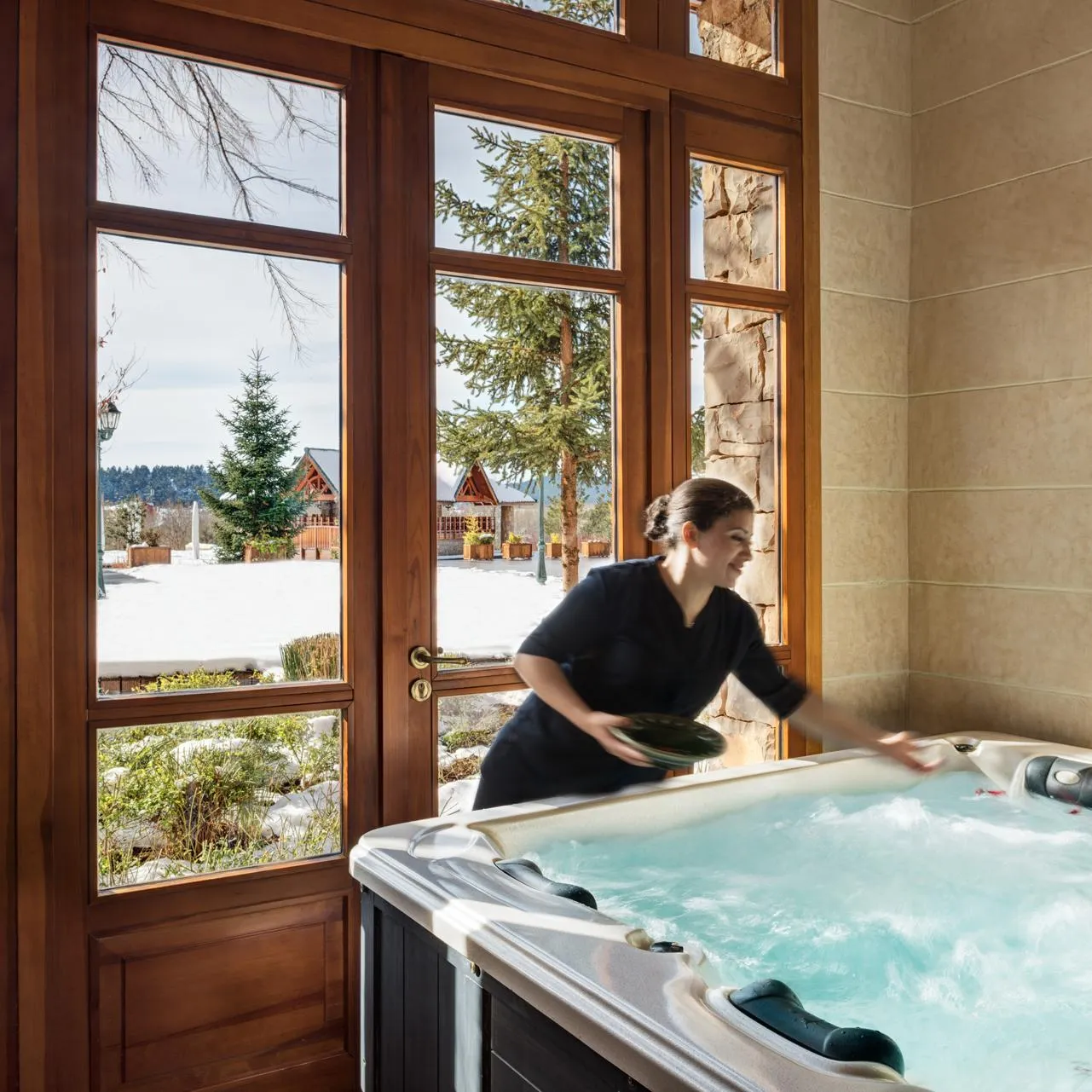 A woman cleans a jacuzzi in a bright room with large windows overlooking a snowy landscape.