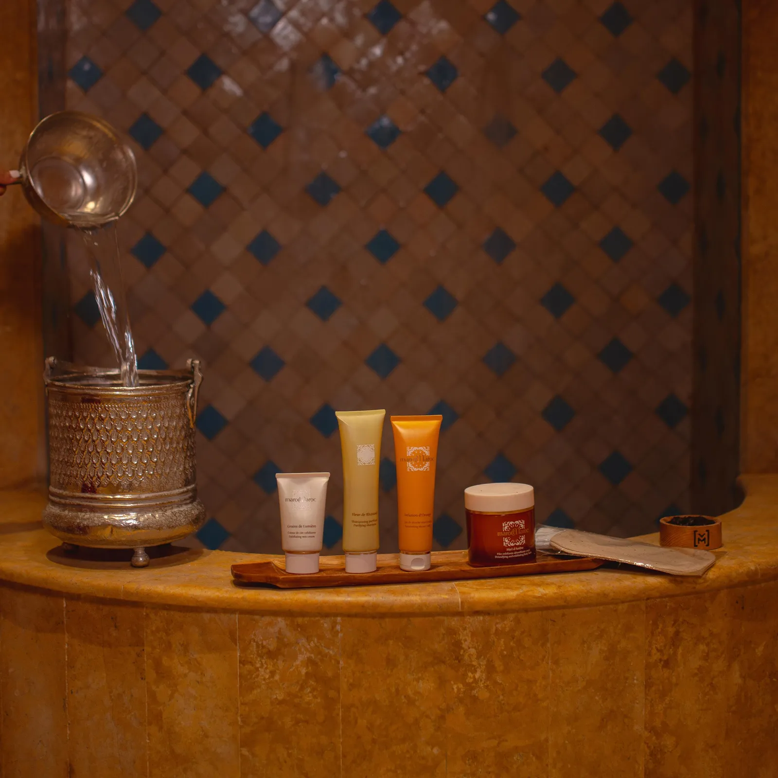A wooden tray with cosmetic product tubes, a jar, and a spatula, next to a metal container with a spout, on a stone shelf with a tiled wall in the background.