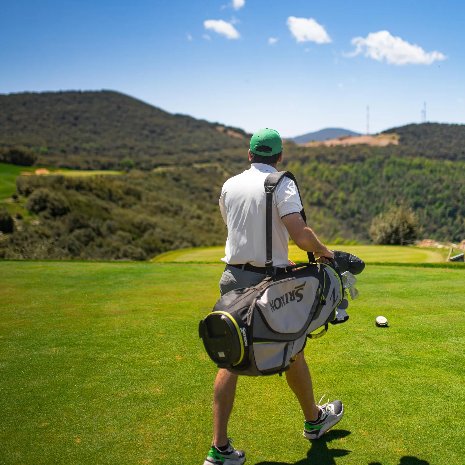 A golfer carrying a bag of clubs walks on a green with hills in the background.