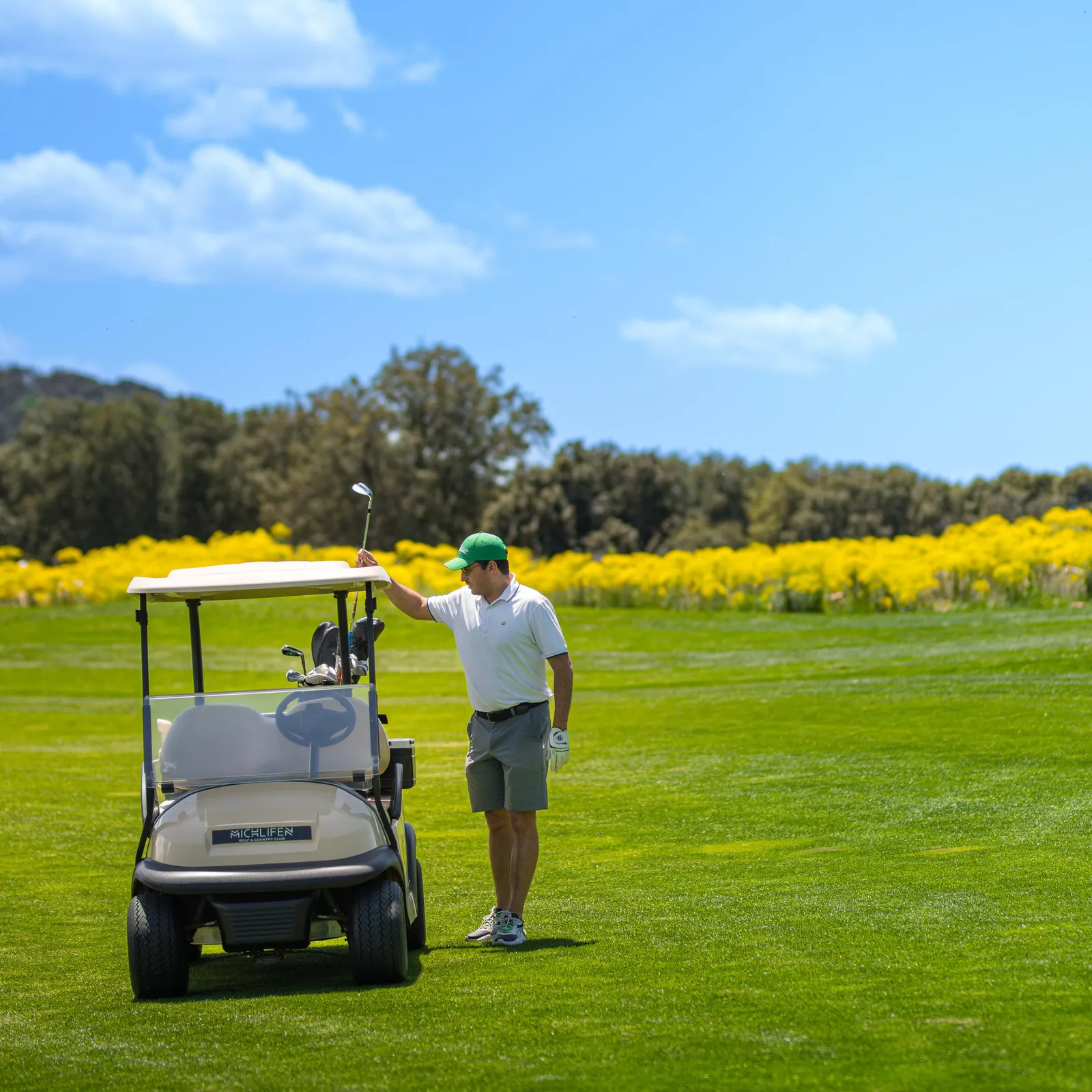 A golfer in green and white attire stands near a golf cart on a golf course with yellow flowers in the background.
