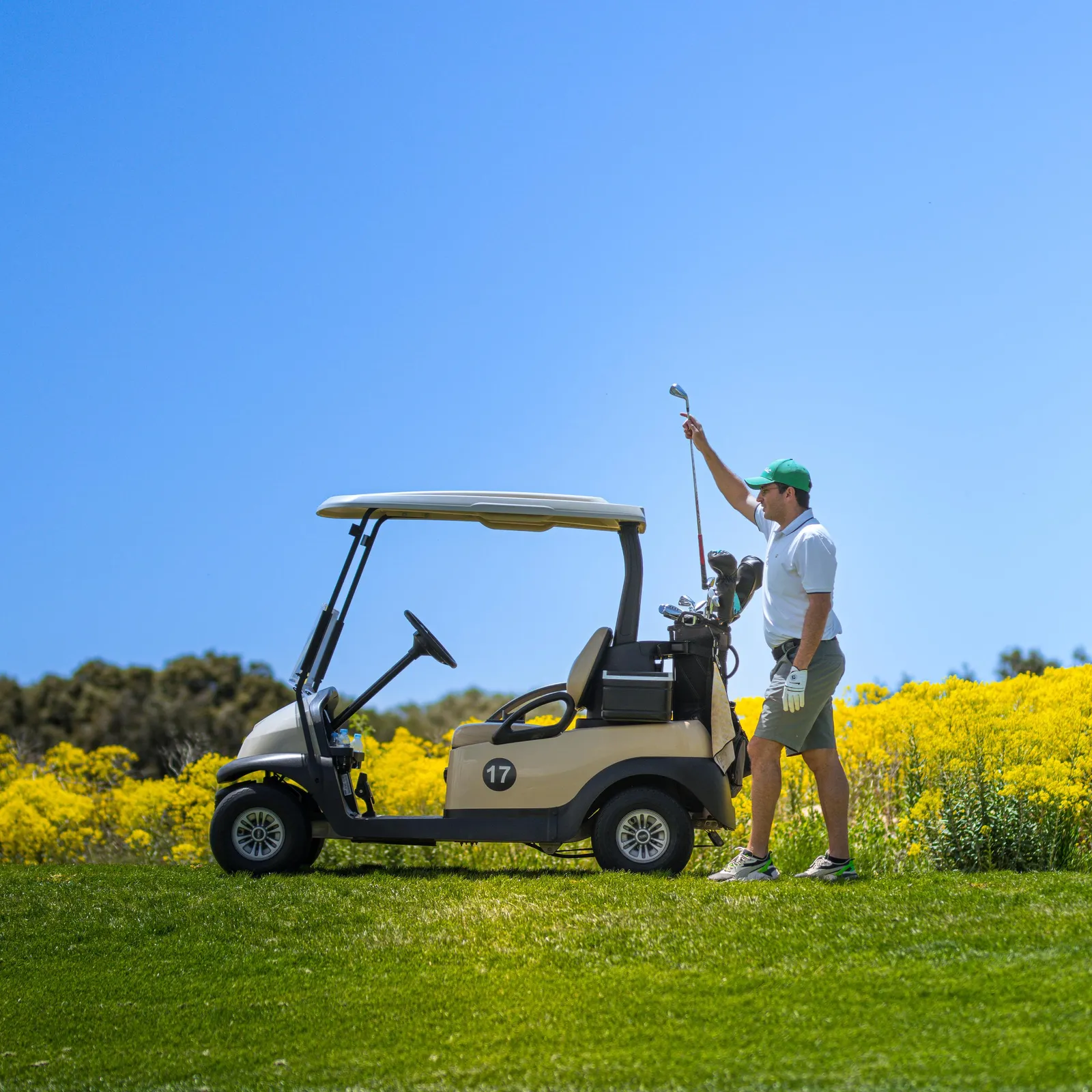 A golfer holds a golf club near a golf cart on a golf course with yellow flowers in the background.