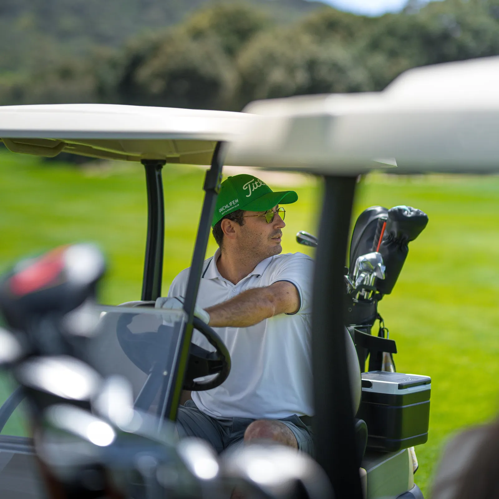 A man in a white shirt and green cap drives a golf cart on a course, with golf clubs in the back.
