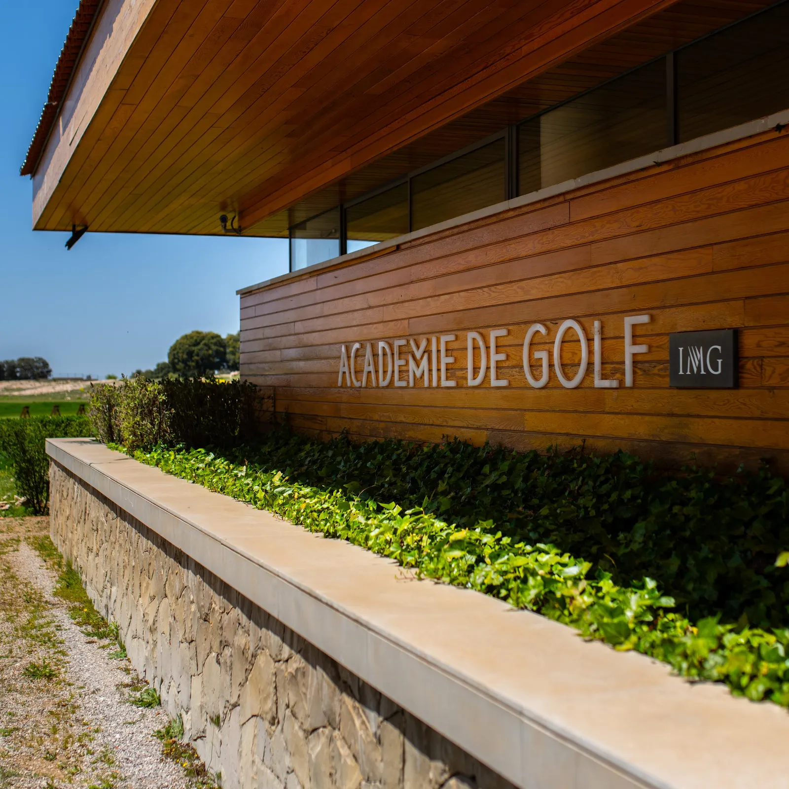 Wooden building with the inscription "IMG Golf Academy" and a stone wall with green plants