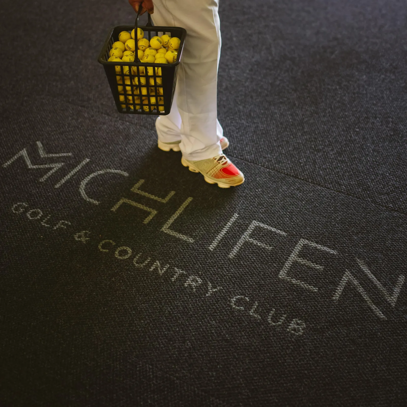 A person is carrying a basket filled with tennis balls on a floor with markings.
