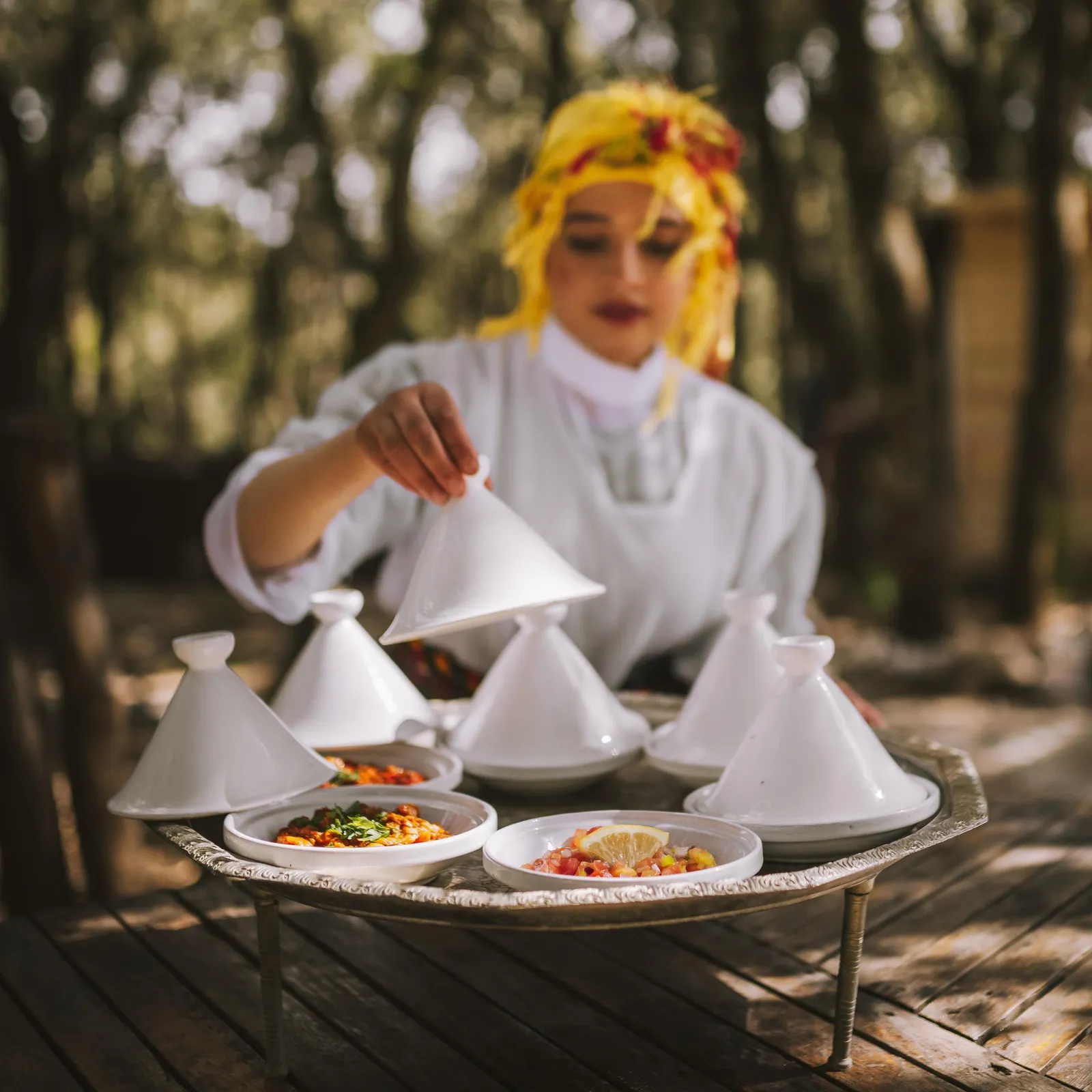 A person in white clothing and a yellow hat prepares dishes in tajines placed on a metal tray on a wooden table outdoors.