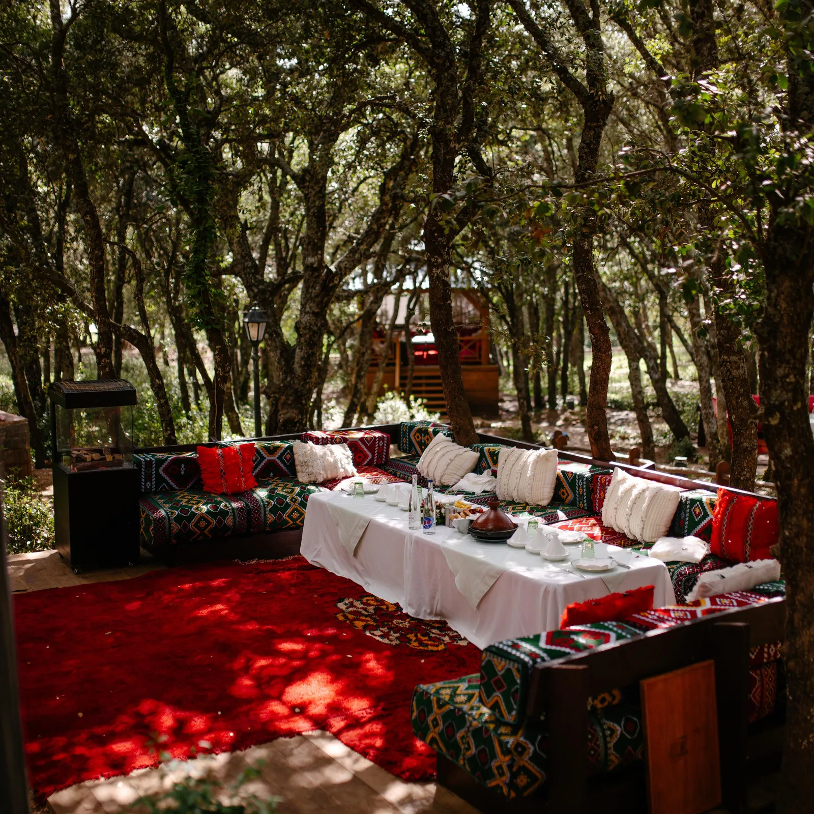 An outdoor space arranged with colorful cushions, a table covered with a white tablecloth, and trees around.