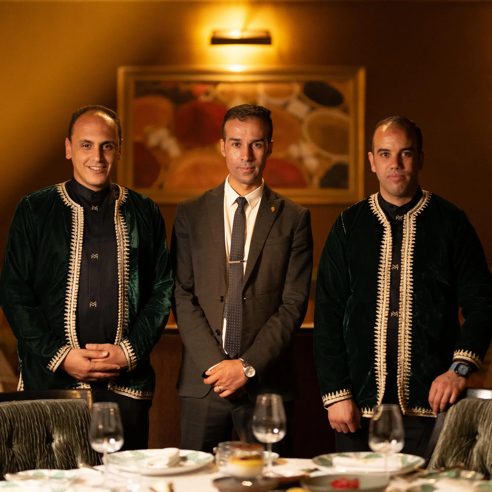 Three men sitting at a table with wine glasses, two wearing ornate jackets and one in a suit