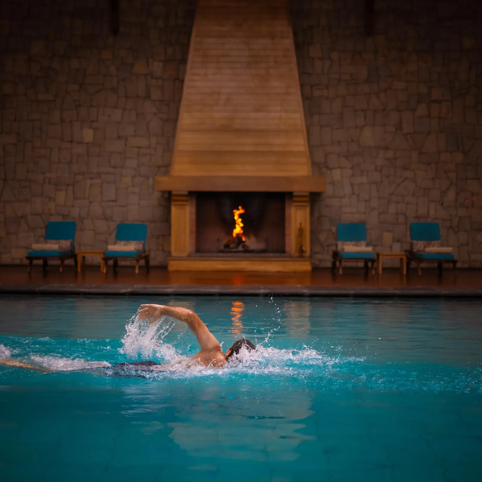 A person is swimming in an indoor pool in front of a lit fireplace.