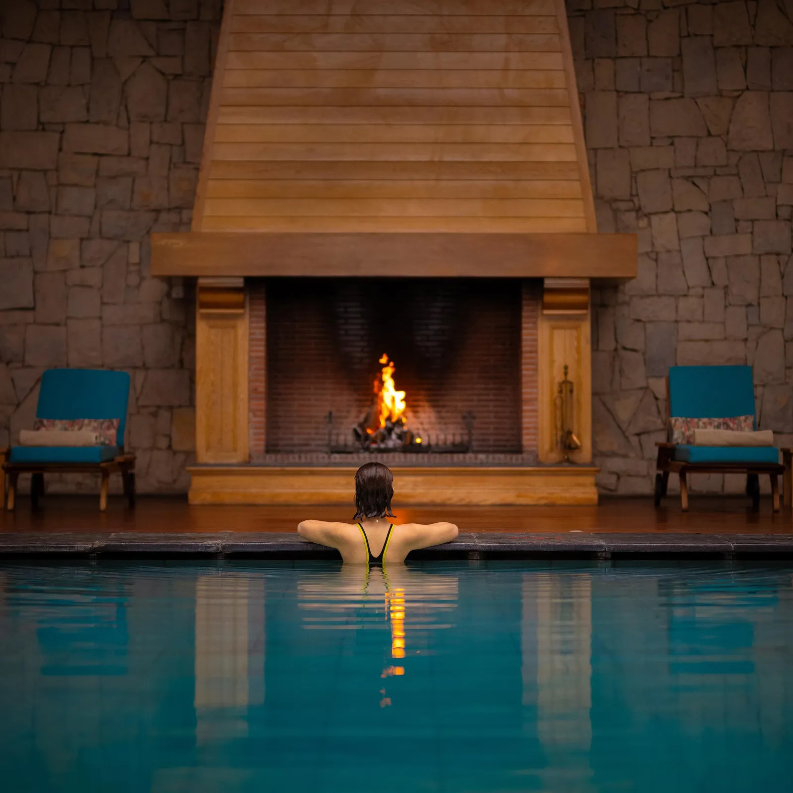 A person is swimming in a pool facing a lit fireplace framed by two blue chairs.