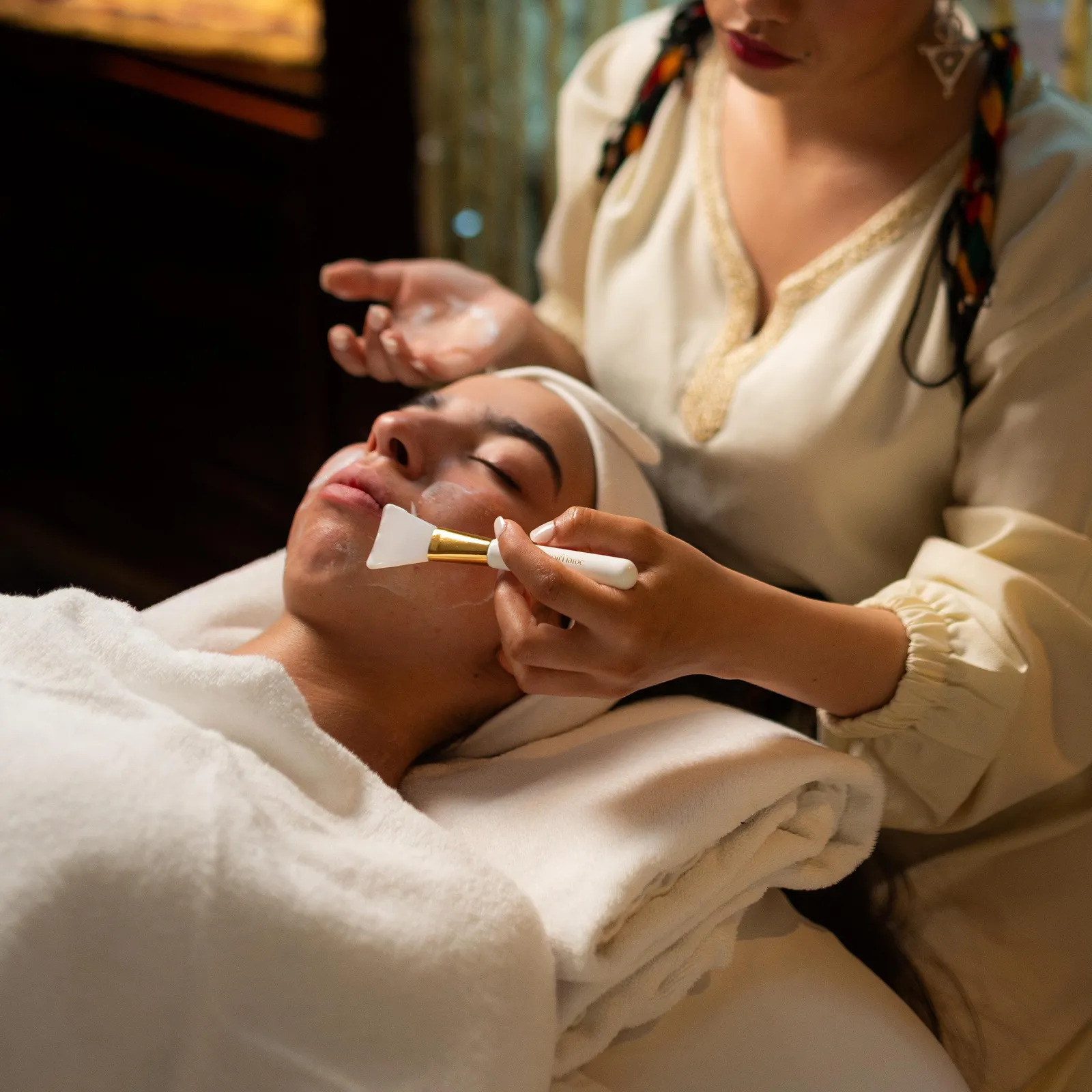 A person applies a mask to the face of another person lying on a towel.