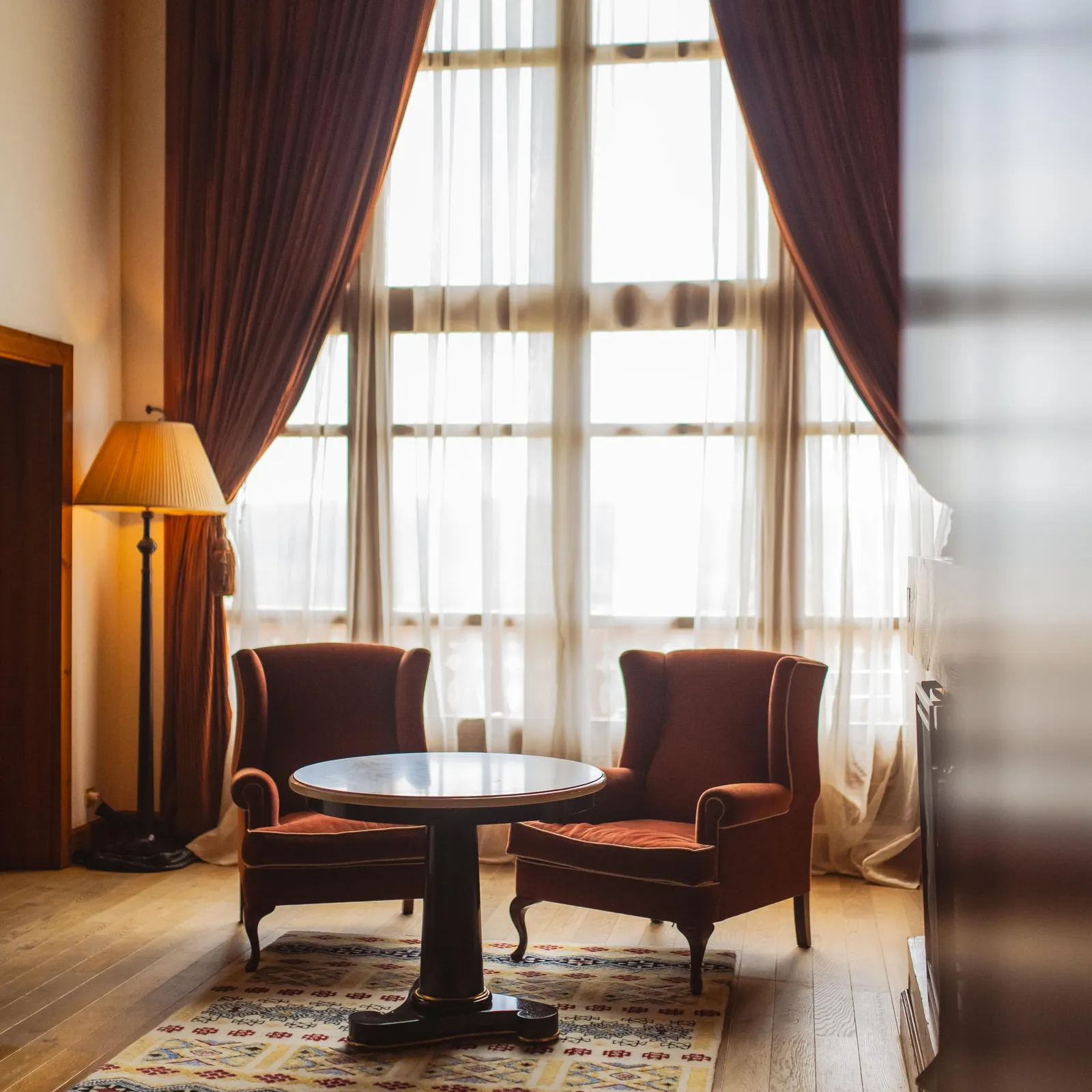 Two red velvet armchairs surround a round table in a living room lit by large windows with brown curtains.