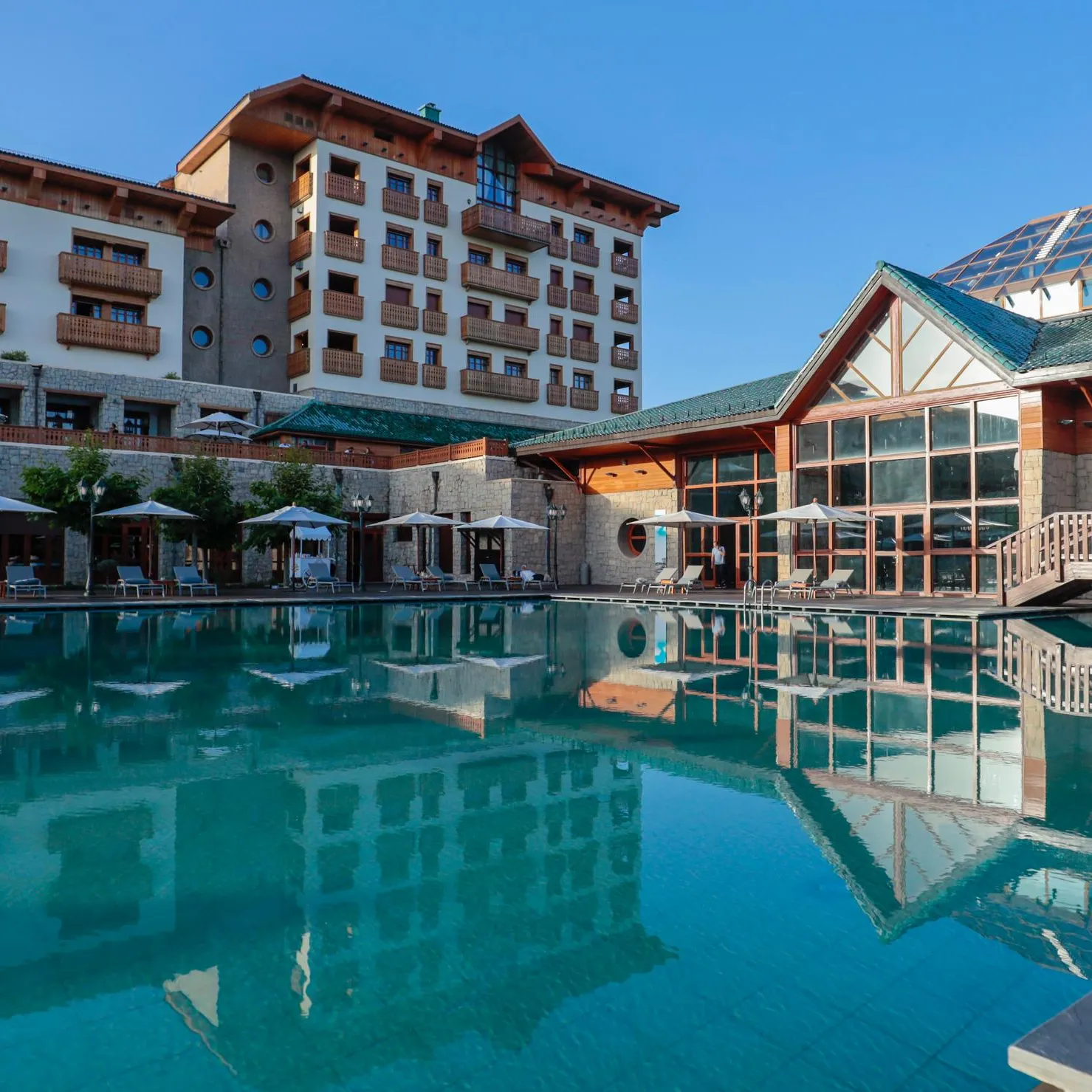 A hotel with an outdoor pool, sun loungers, and umbrellas under a clear sky.