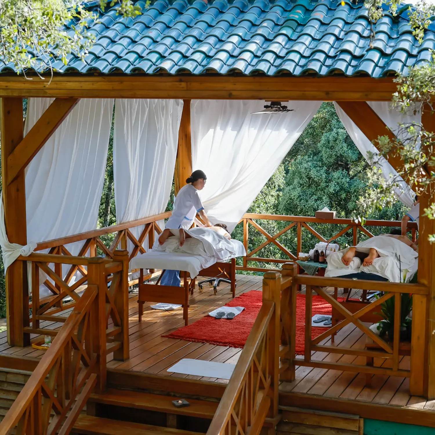 Two people lying on outdoor massage tables under a wooden awning with a blue-tiled roof, surrounded by trees.