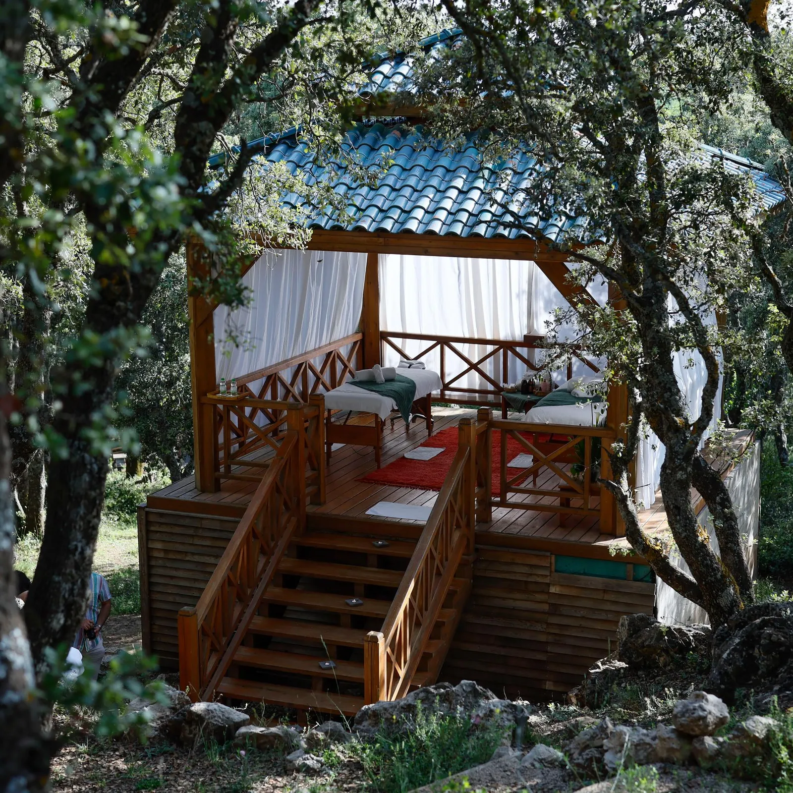 A wooden cabin with a bed and cushions is located in a forest, surrounded by trees and accessible via a staircase.