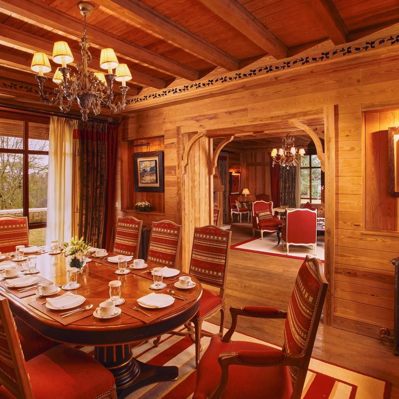 Wooden dining room with a large round table, red chairs, and candelabras, overlooking a garden through large windows.