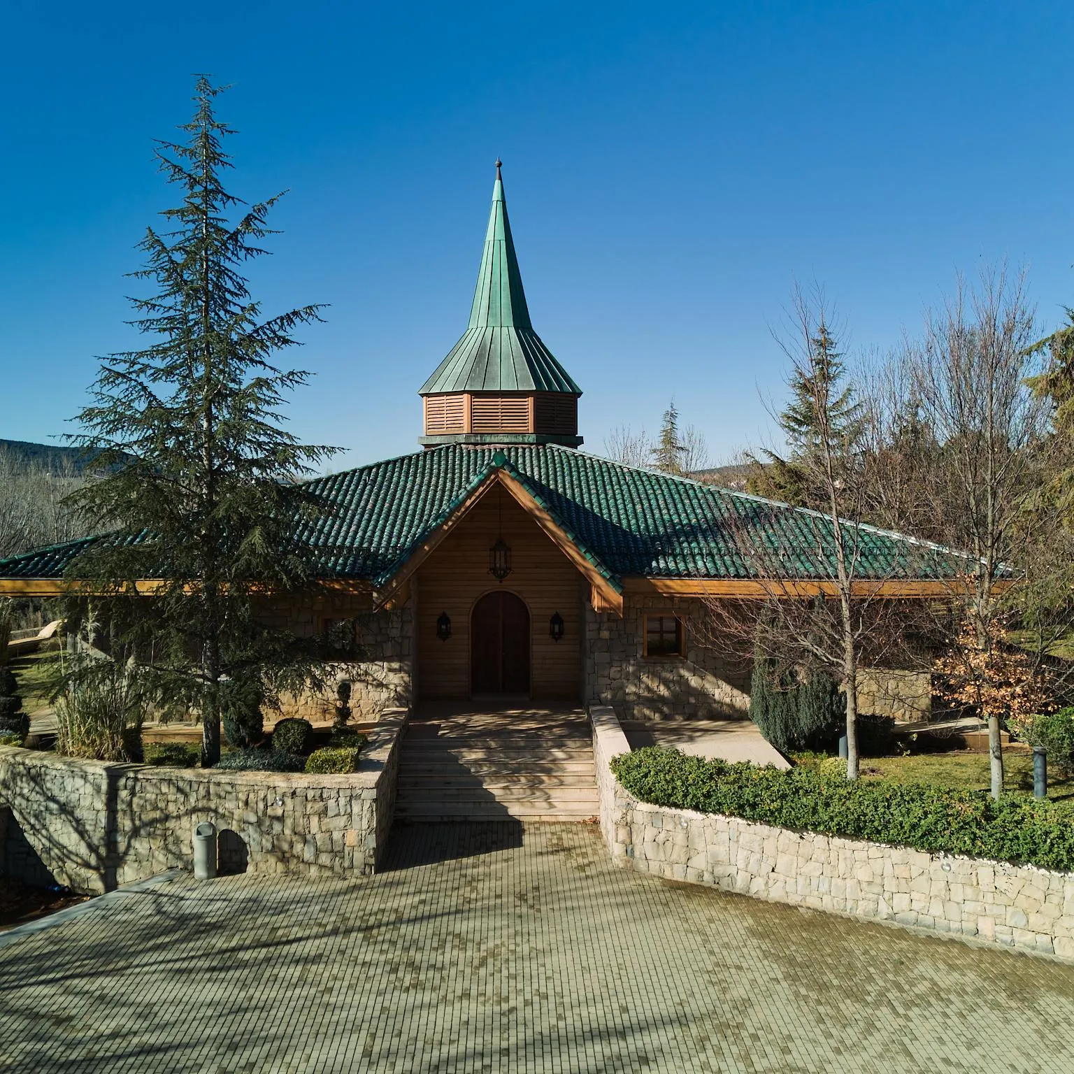 Small wooden church with a green roof and a pointed spire, surrounded by trees and shrubs, located in a well-maintained garden with stone walls and paved paths.