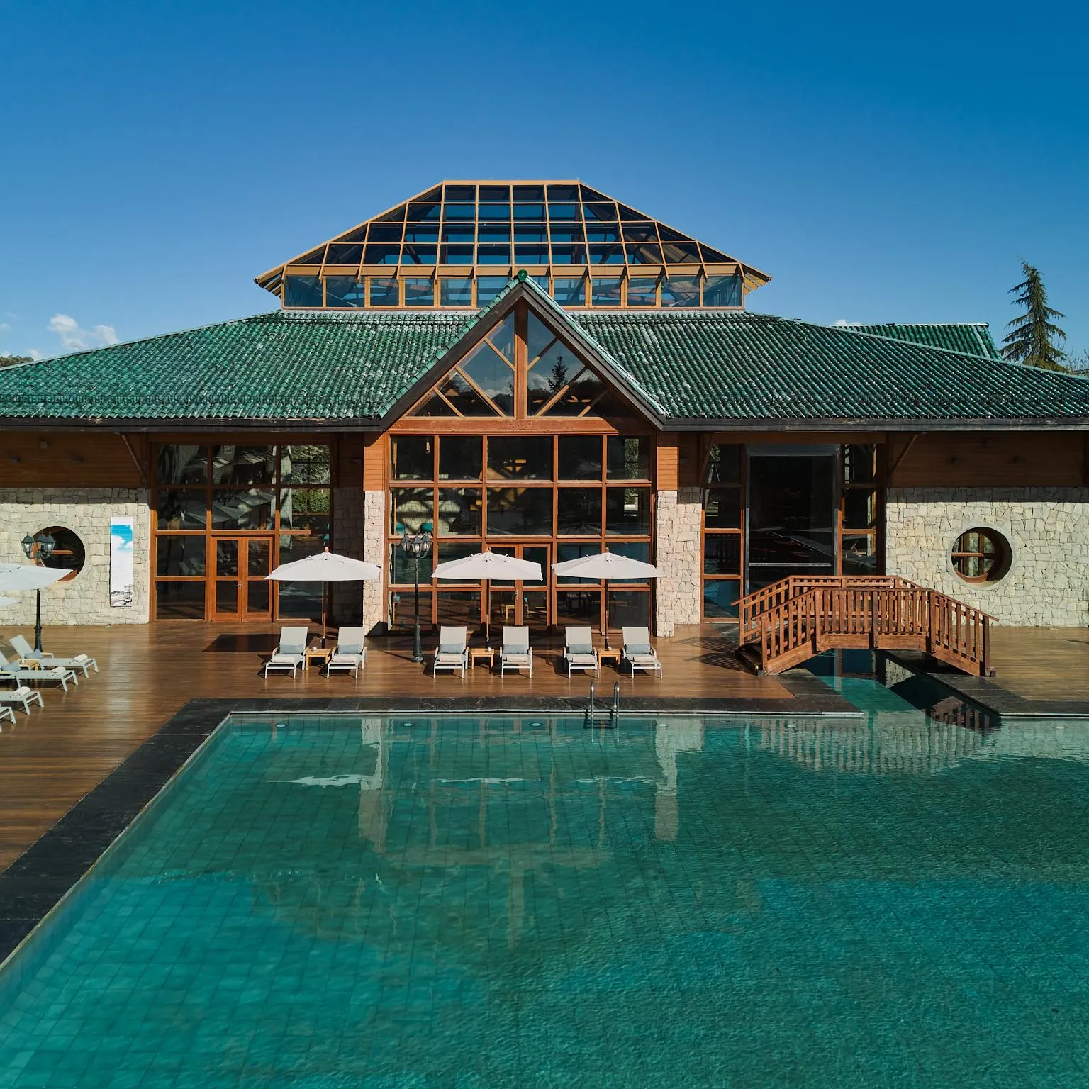 A stone and wood building with a green roof and a glass extension, in front of an outdoor pool surrounded by lounge chairs and umbrellas.