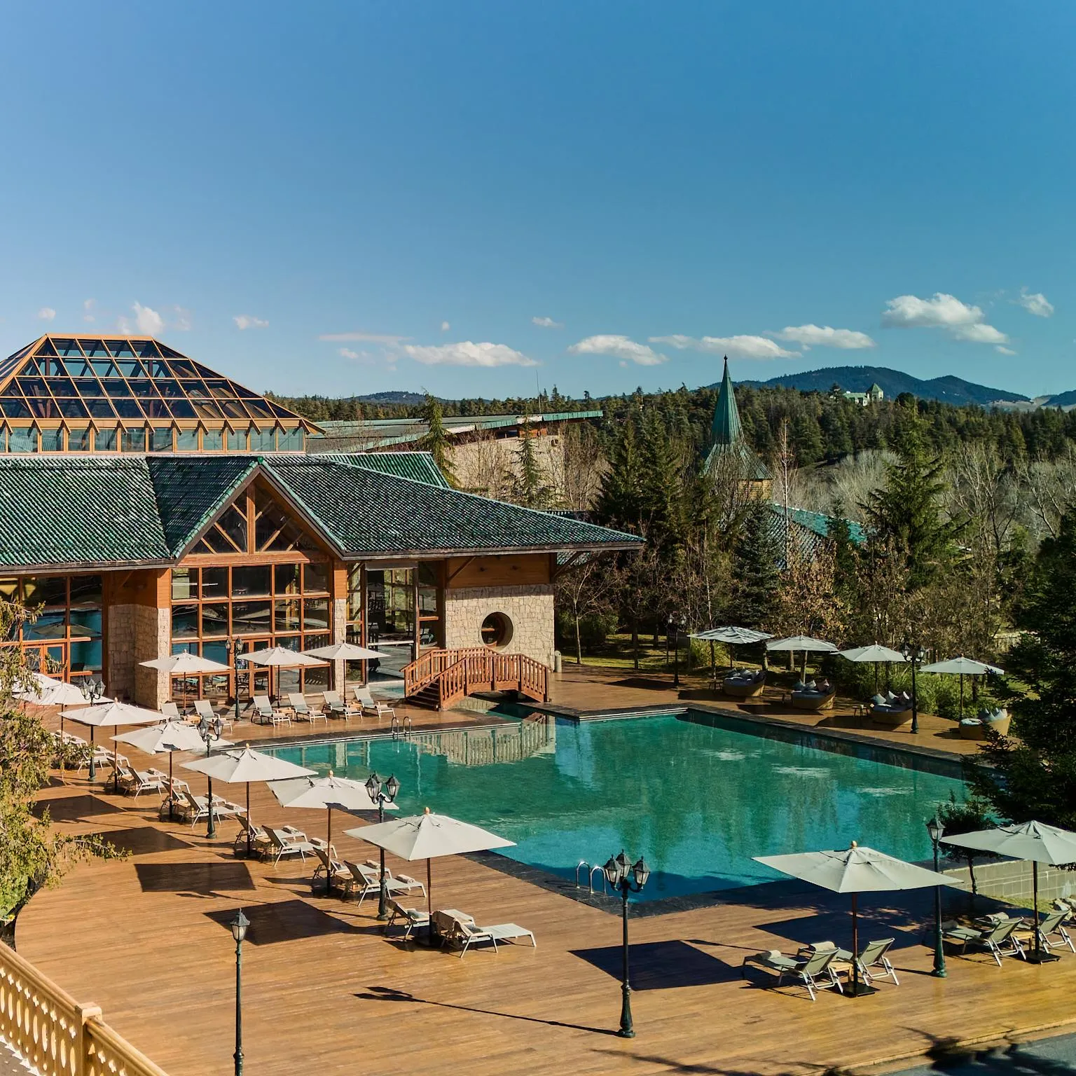 Outdoor swimming pool surrounded by sun loungers and parasols, with a wooden building and green roof in the background, in a mountainous natural setting.