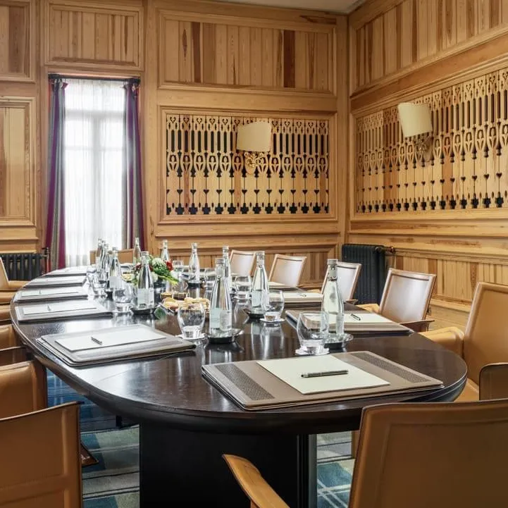 Meeting room with an oval table surrounded by leather chairs, glasses and notepads on the table, wooden walls with carved patterns and windows with curtains.