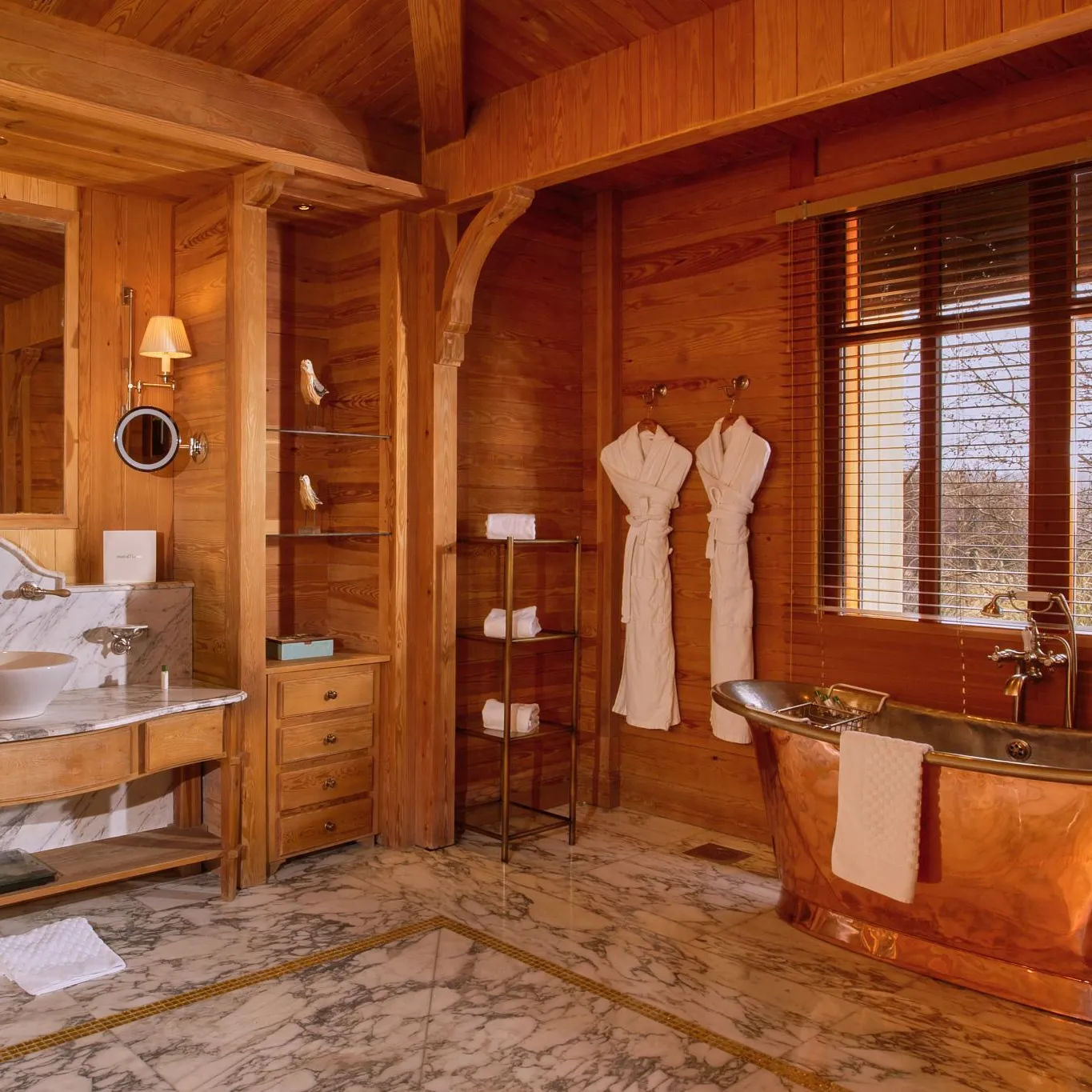 Wooden bathroom with marble sink, copper bathtub, and two hanging robes near a window.