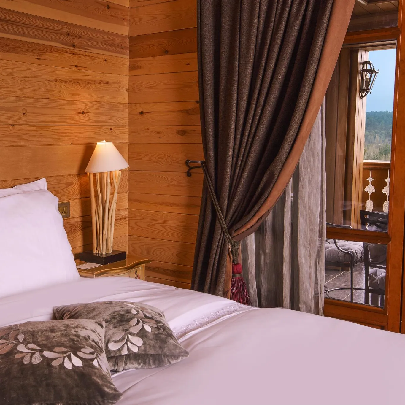 Wooden room with a bed, cushions, a bedside lamp, and a French window opening onto a balcony with a view of a landscape.
