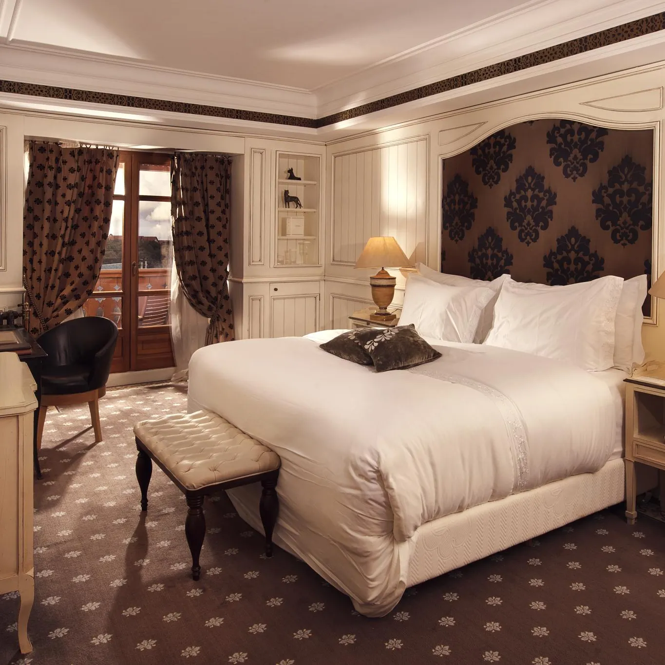 Elegant room with a large white bed, bedside tables, a desk, a chair, and a French door leading to a balcony.