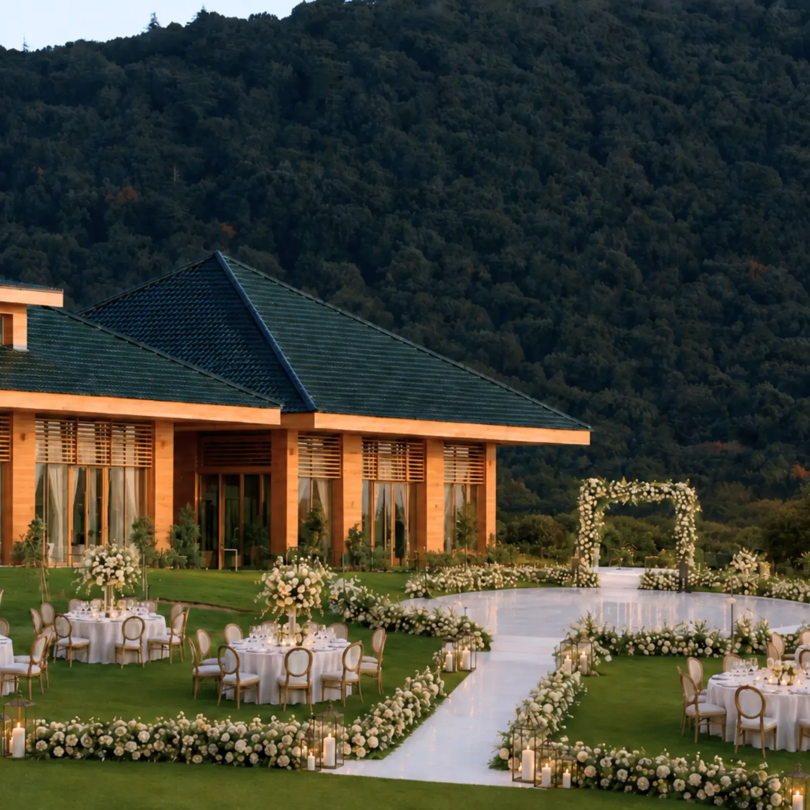 An outdoor space arranged for an event with tables, chairs, and floral decorations, in front of a building with orange walls and a green roof, surrounded by a wooded hill