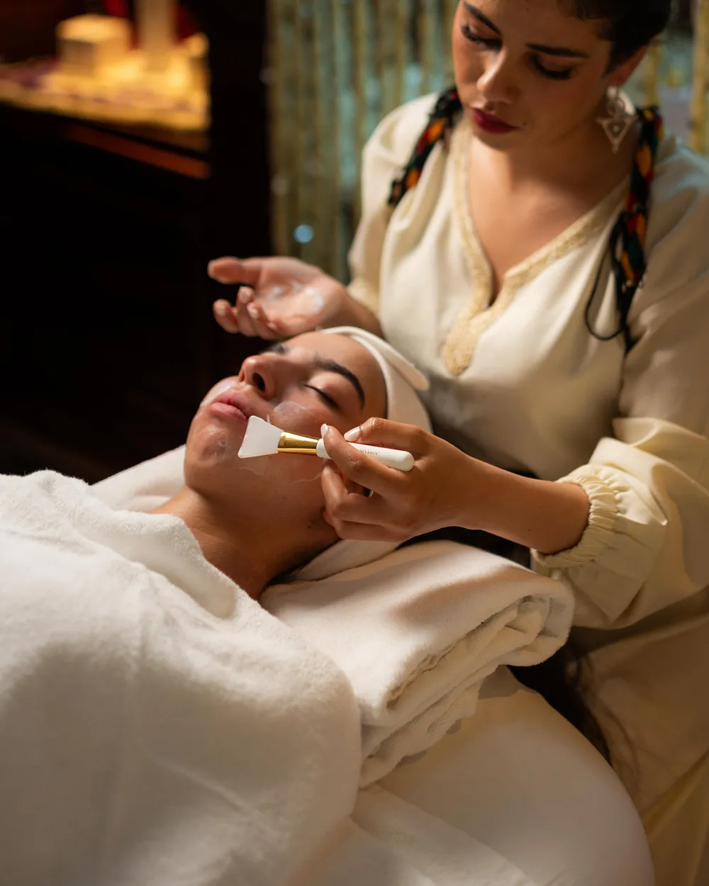 A person applies a mask to the face of another person lying on a towel.