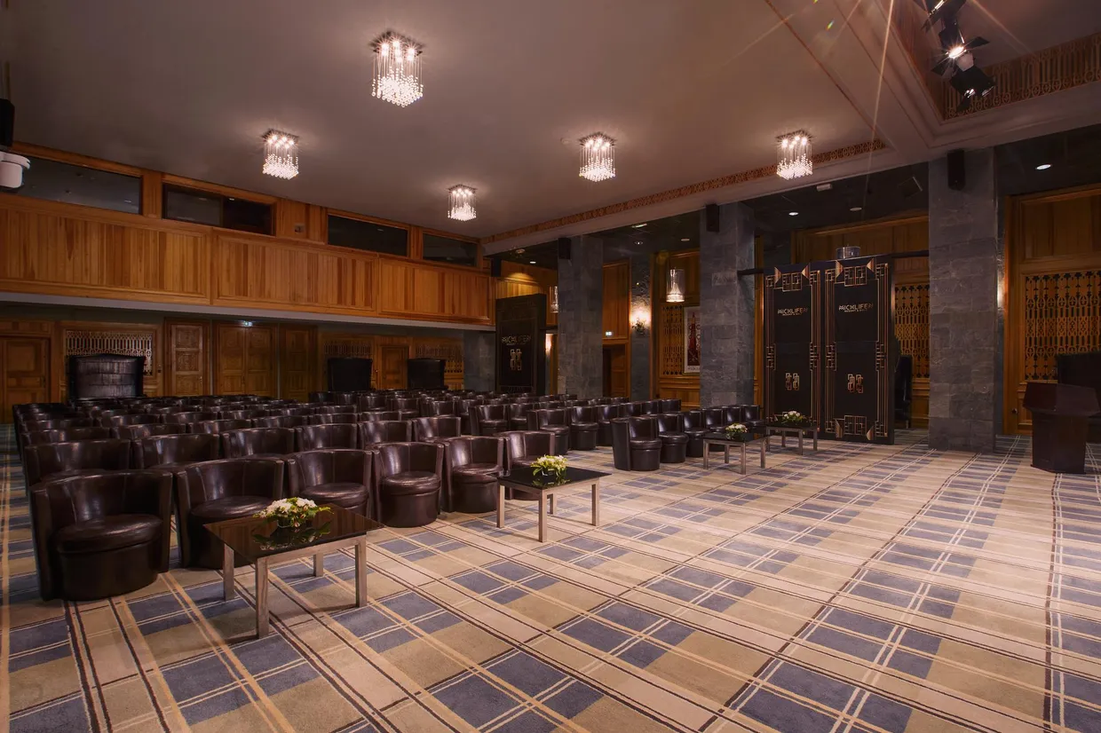 Conference room with rows of brown leather armchairs, low tables with flowers, wooden walls, and chandeliers on the ceiling.