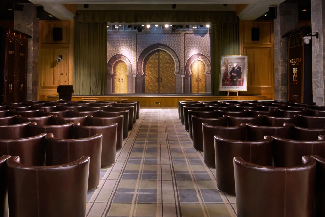 Conference room with leather seats arranged in rows facing a stage adorned with architectural motifs and lit, with a portrait on an easel to the right.