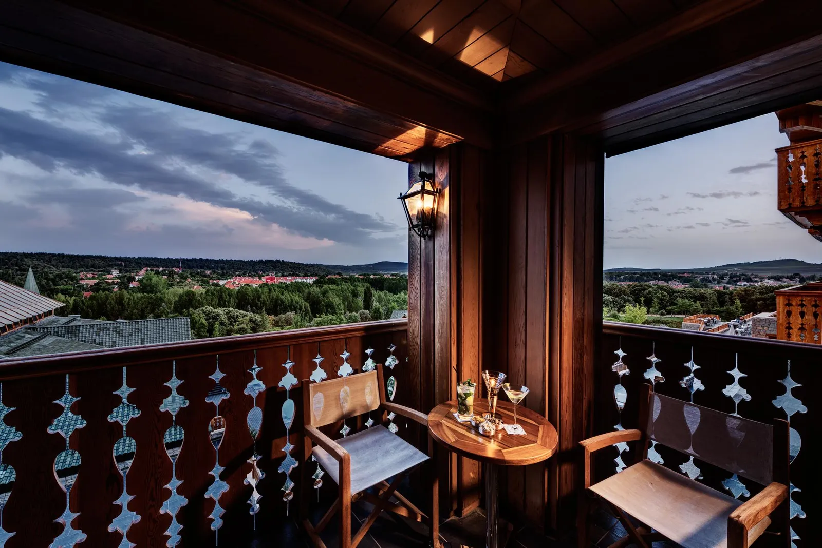 Wooden balcony with table, chairs, and glasses, offering a panoramic view of a city and hills under a cloudy sky.