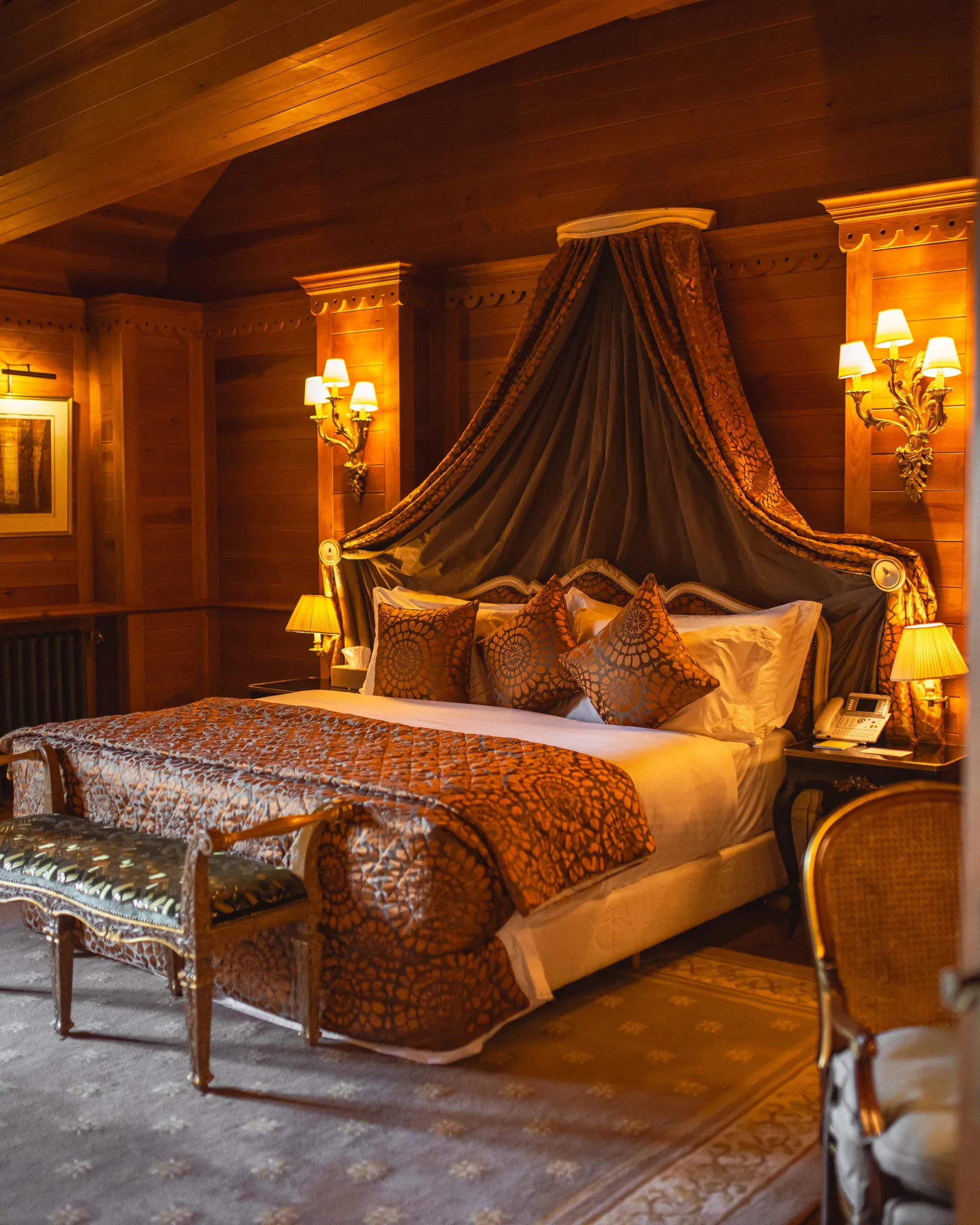 Luxurious room with a large four-poster bed adorned with curtains and cushions, framed by wood paneling and lit by wall lamps and bedside lamps.
