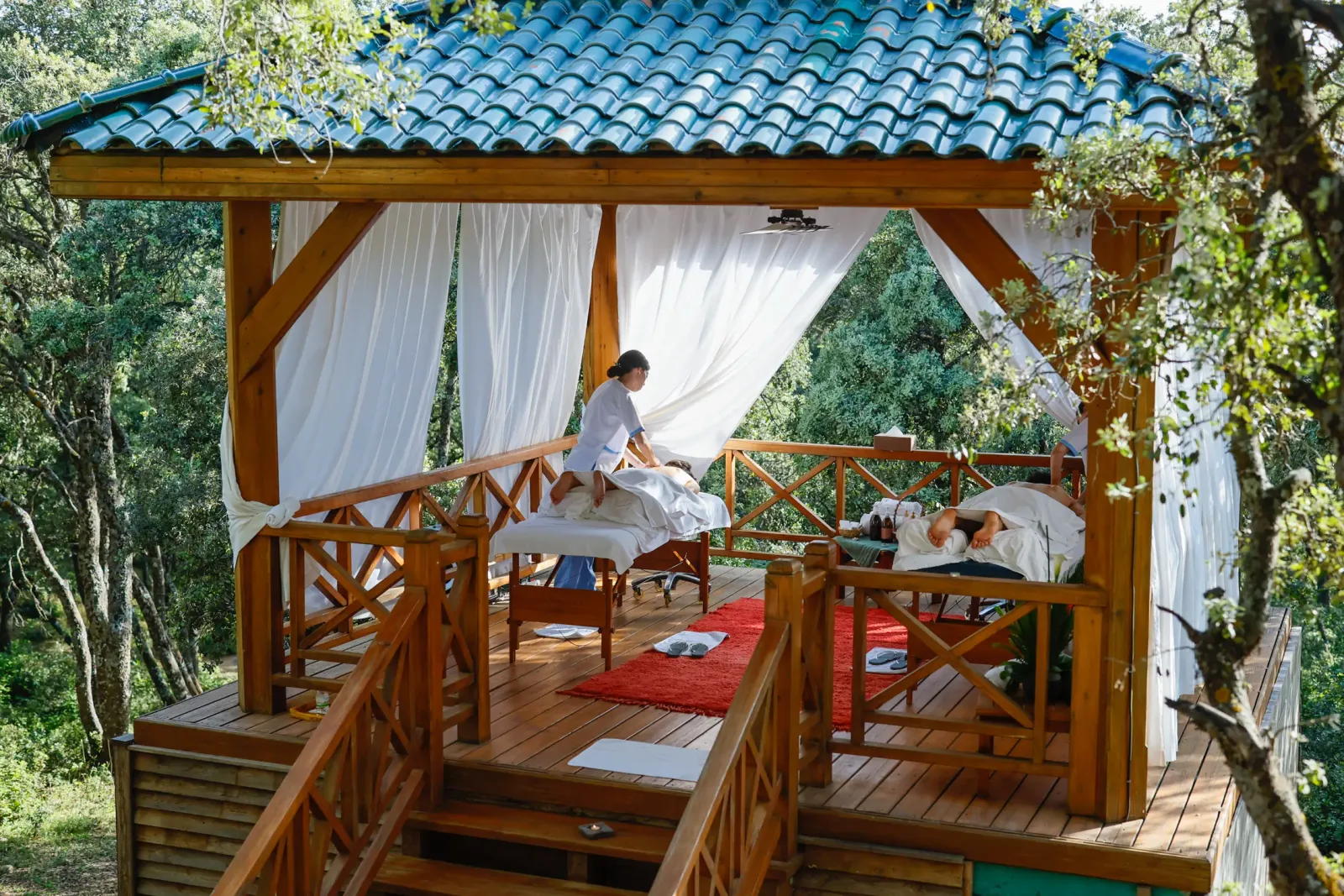 Deux personnes allongées sur des tables de massage en plein air sous un auvent en bois avec un toit en tuiles bleues, entourées d'arbres