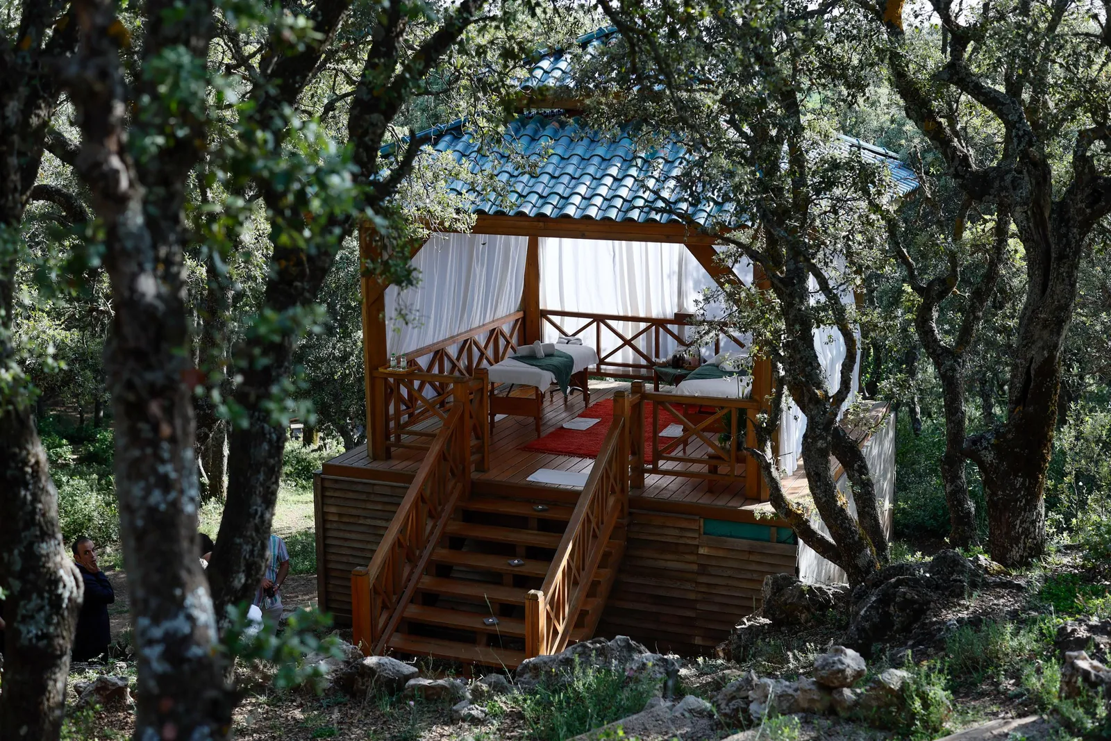 A wooden cabin with a bed and cushions is located in a forest, surrounded by trees and accessible via a staircase.