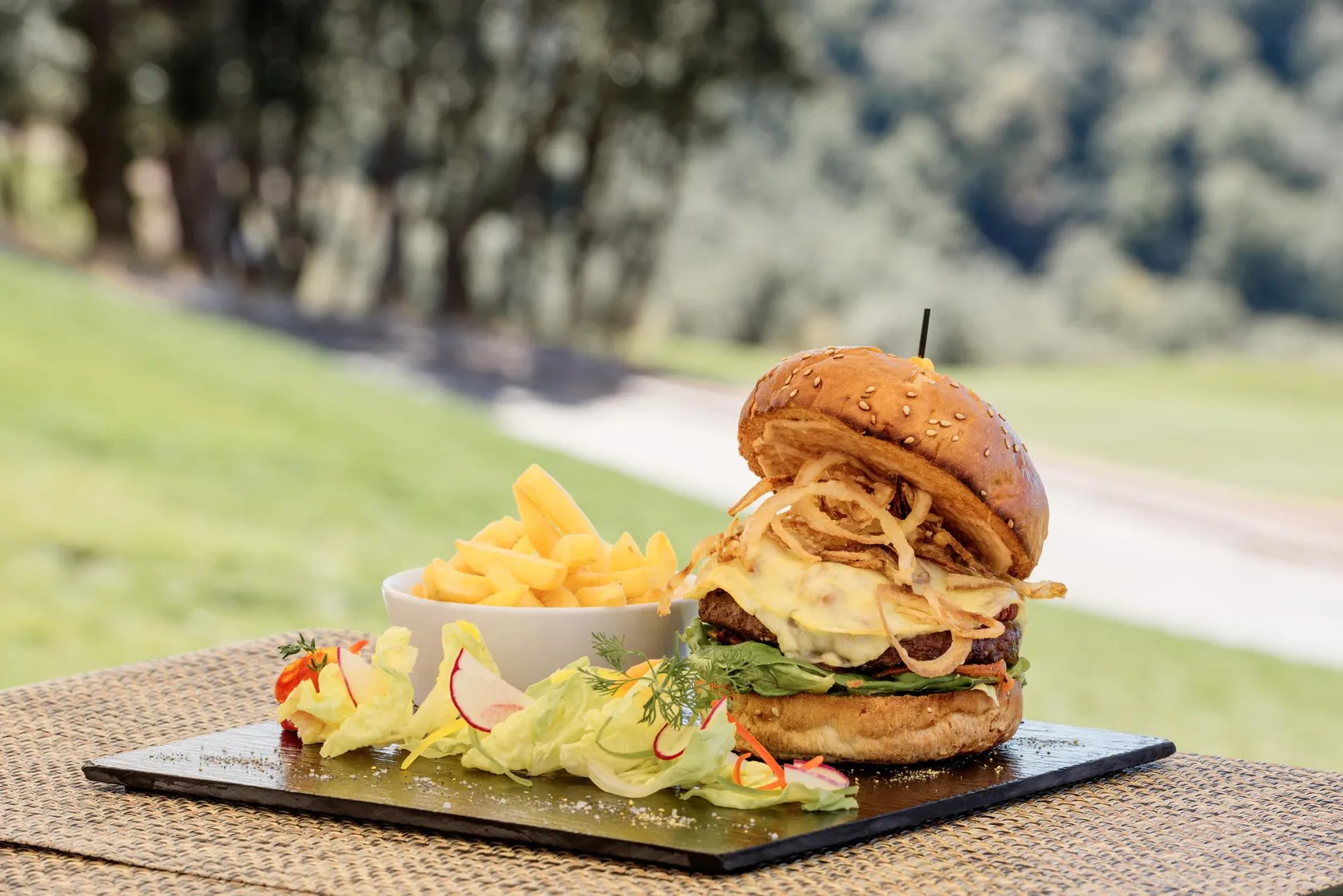 A burger with fried onions and lettuce, served with fries in a bowl, placed on a black rectangular plate.