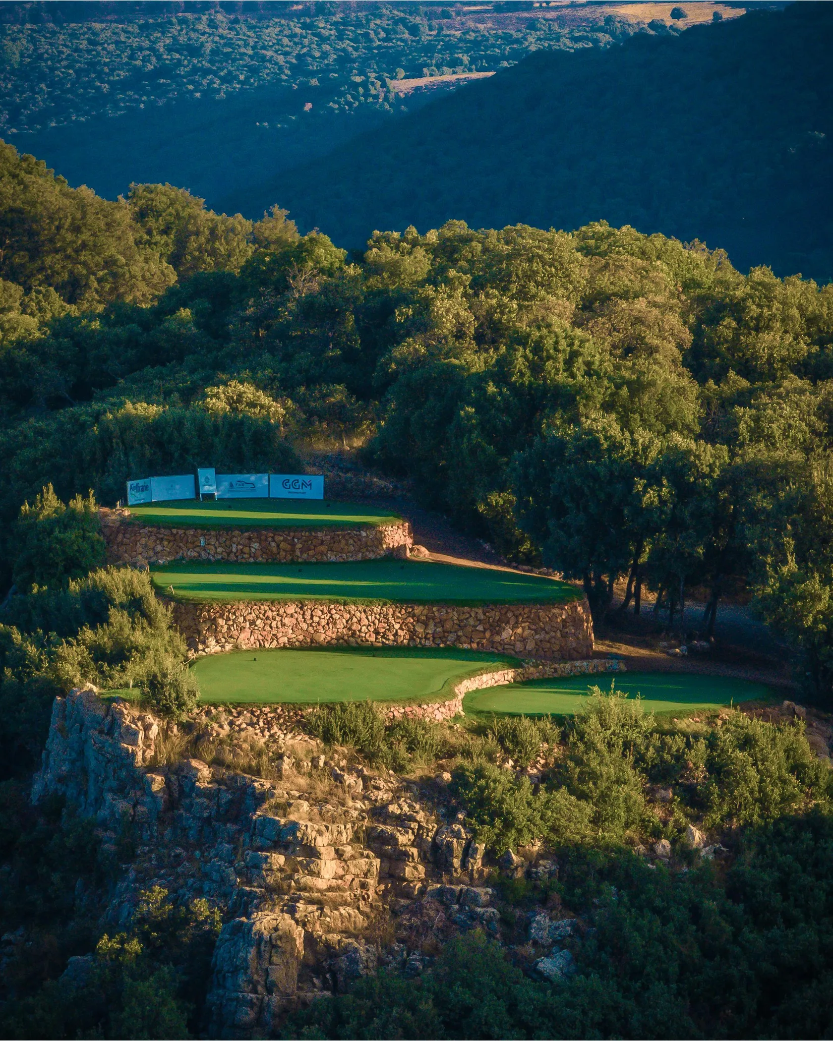 Golf on a terraced course with stone walls, surrounded by trees and vegetation, with a view of a valley in the background