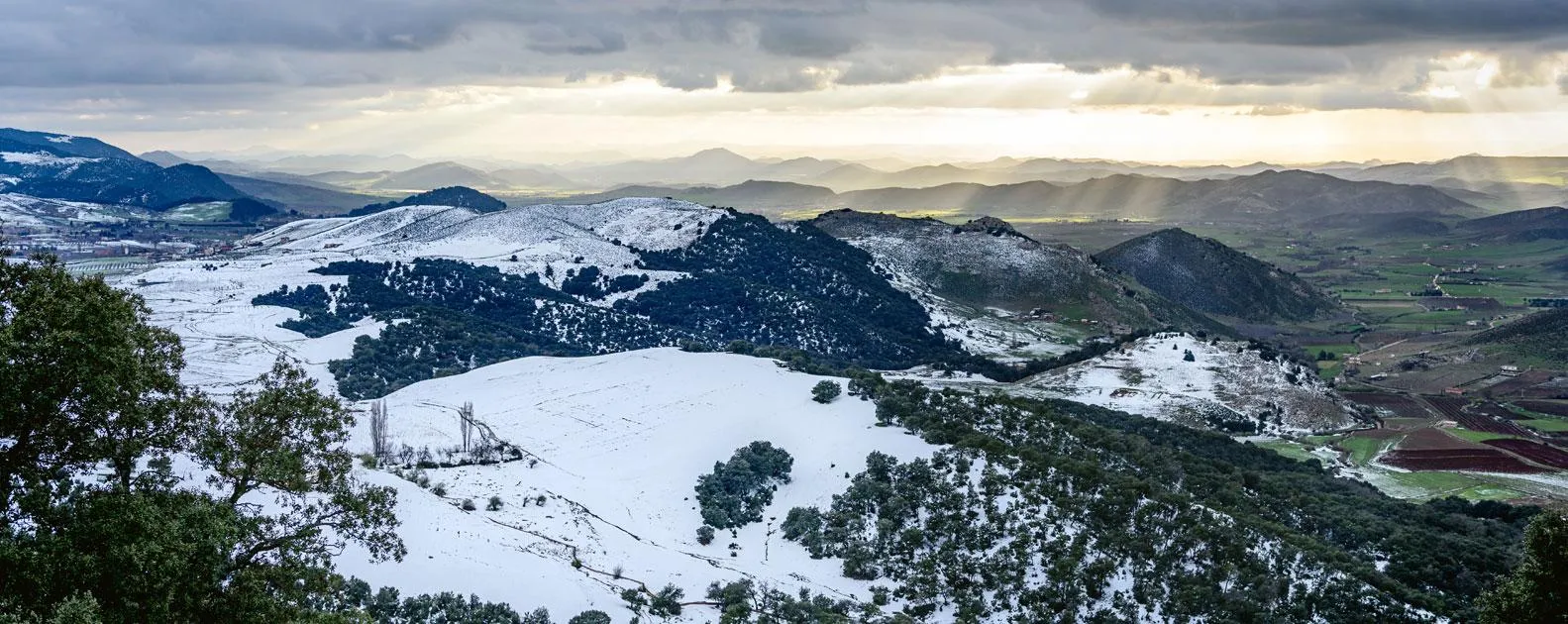 Snowy mountainous landscape with hills, fields, and forests under a cloudy and sunny sky