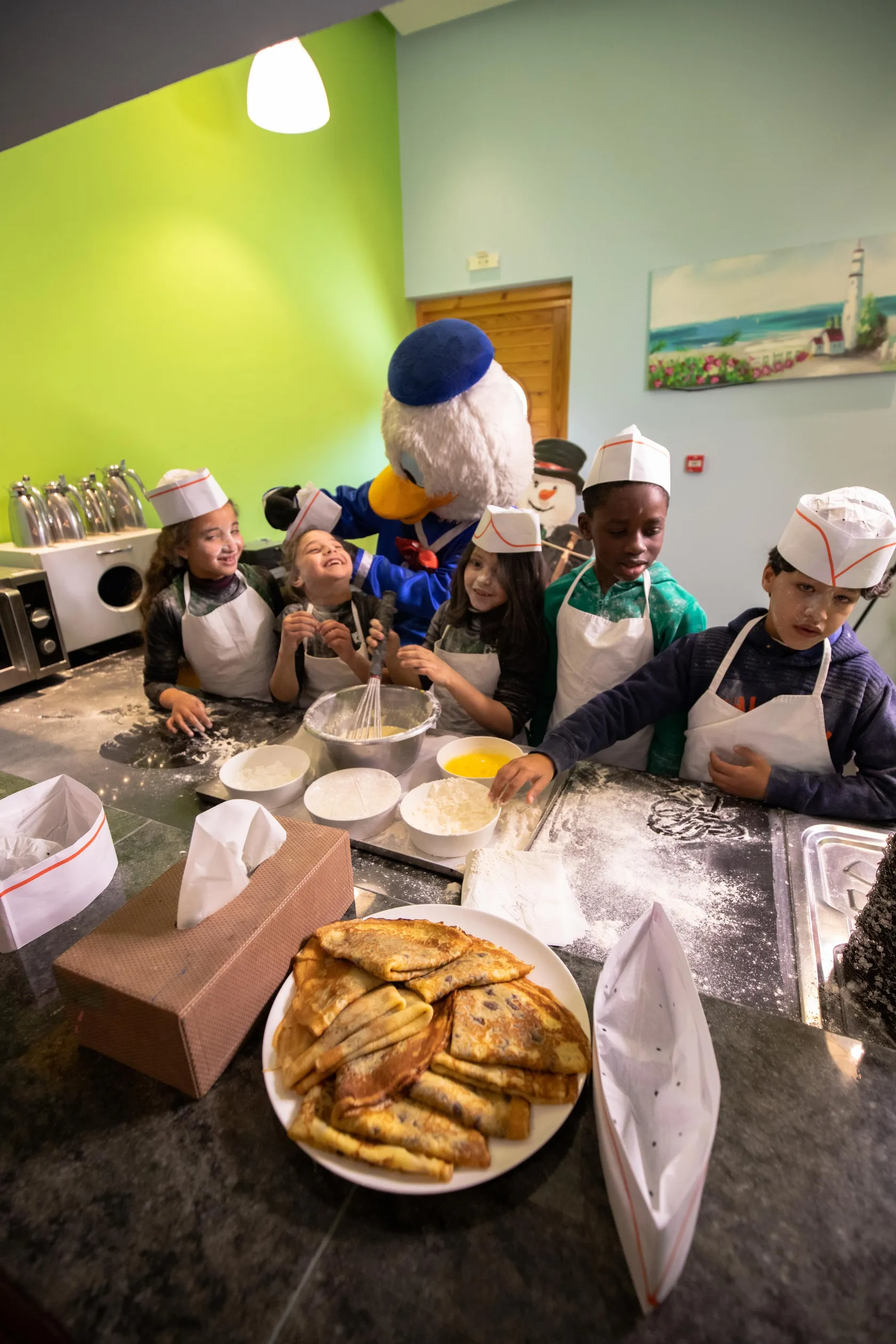 A group of children and a mascot character cook together in a kitchen, surrounded by ingredients and baking utensils.