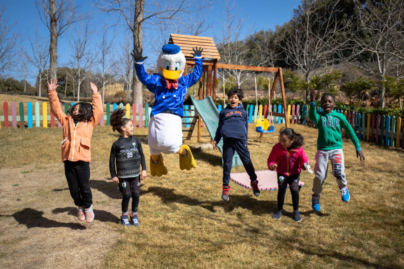 A costumed duck character jumps with five children in a park with wooden games and a colorful fence in the background.