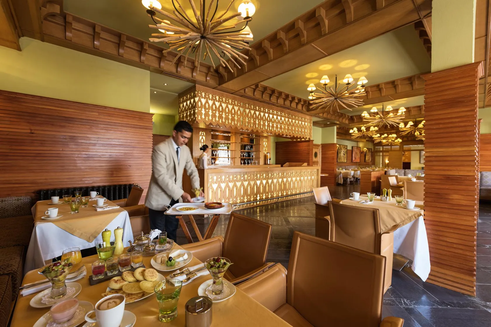 A waiter in a white jacket carries a tray with dishes in an elegant restaurant with wooden walls and decorative chandeliers.