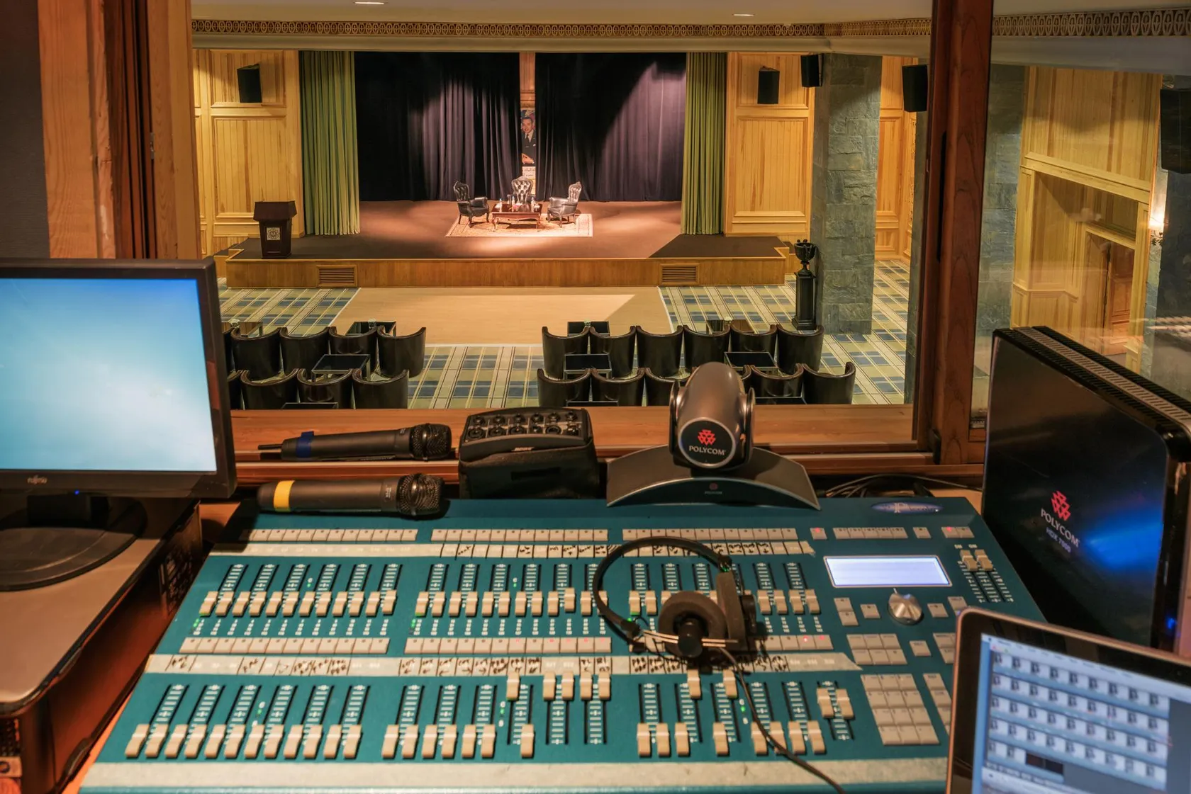Sound mixing desk with screens and microphones in front of a theater stage with chairs and a closed curtain