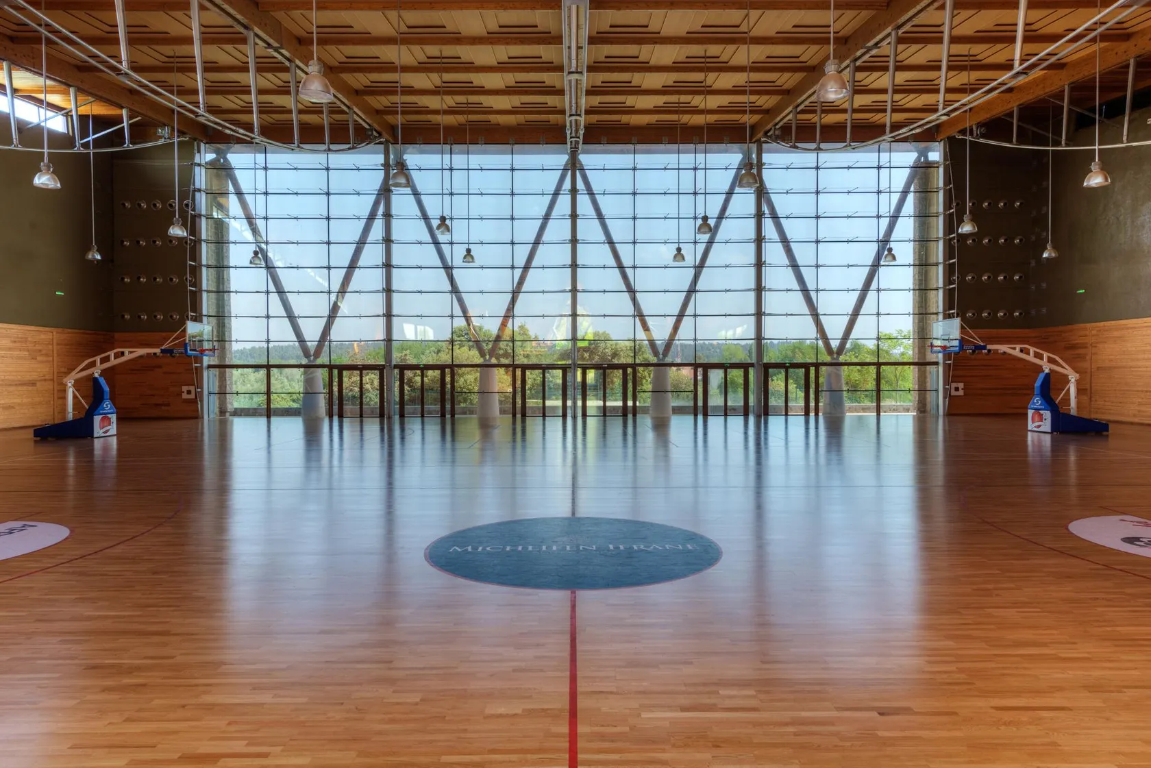 Indoor gymnasium with a polished wooden floor, basketball panels, and a large glass wall facing it
