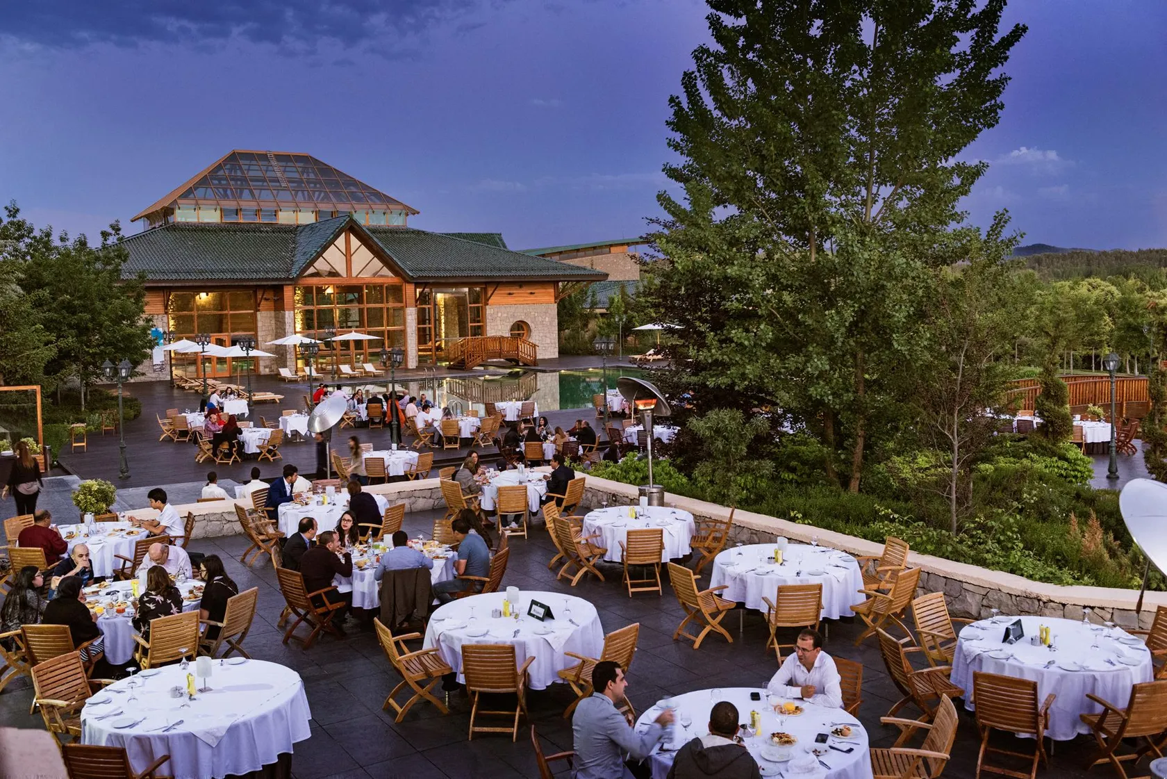 An outdoor restaurant terrace with many tables and chairs, diners eating, and a wooden and stone building in the background.