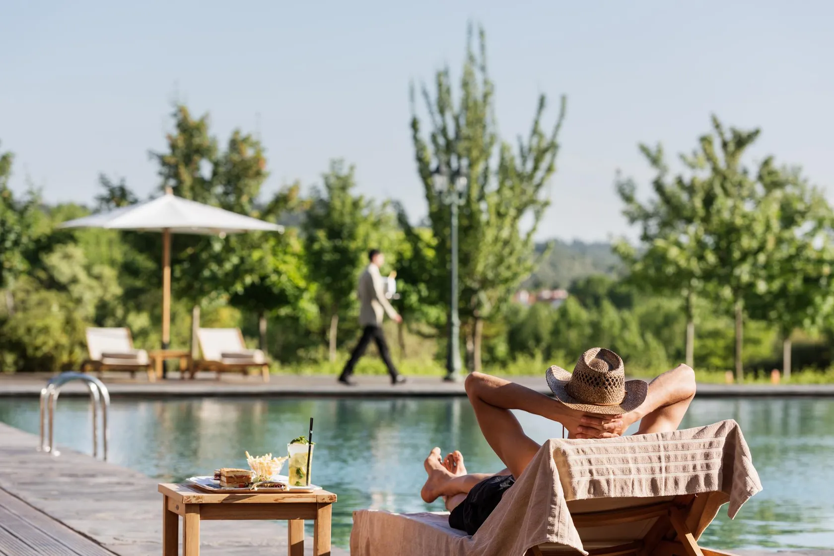 A person in a swimsuit and hat rests on a sun lounger by the poolside, with a low table containing drinks and food nearby.