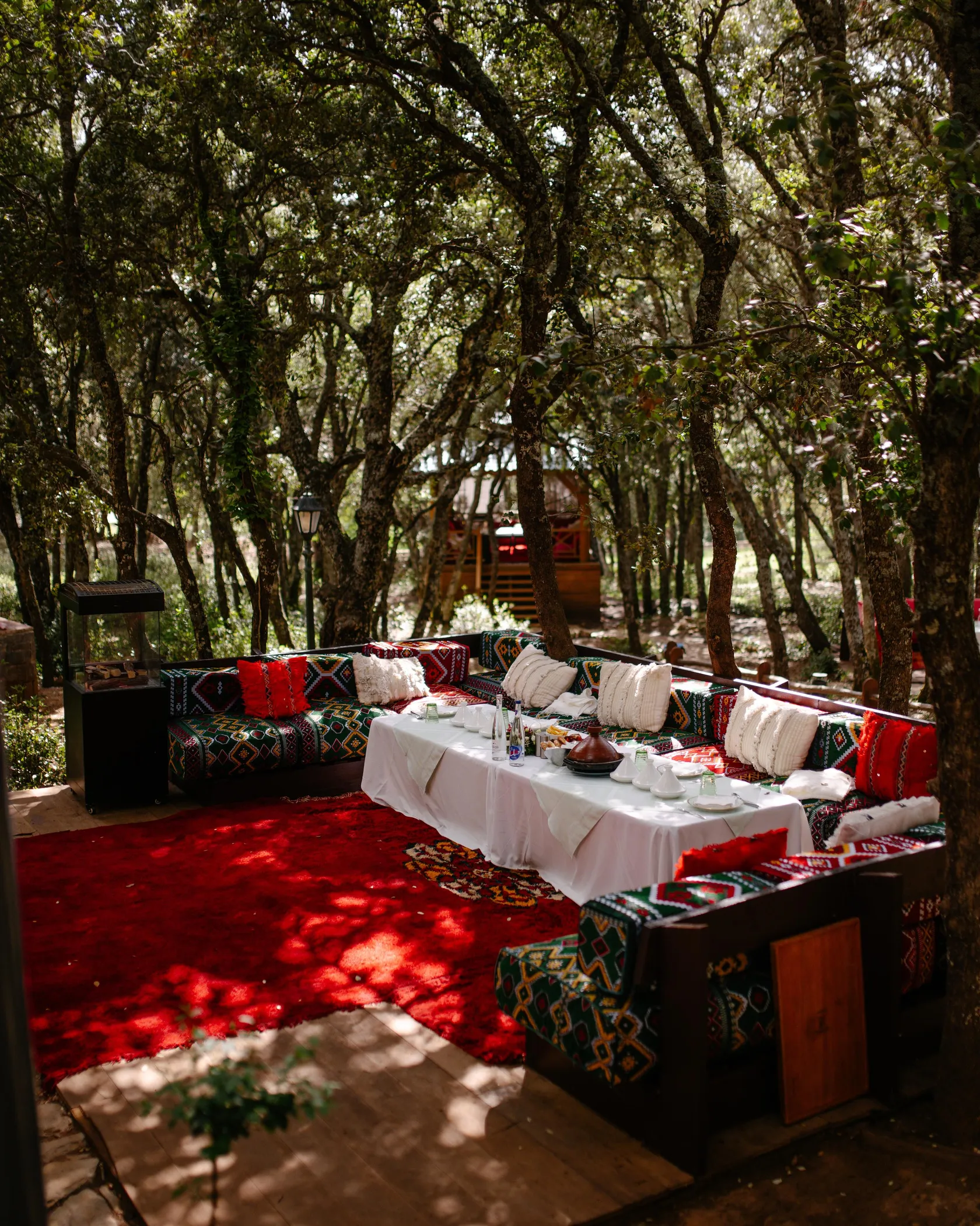 An outdoor space arranged with colorful cushions, a table covered with a white tablecloth, and trees around.