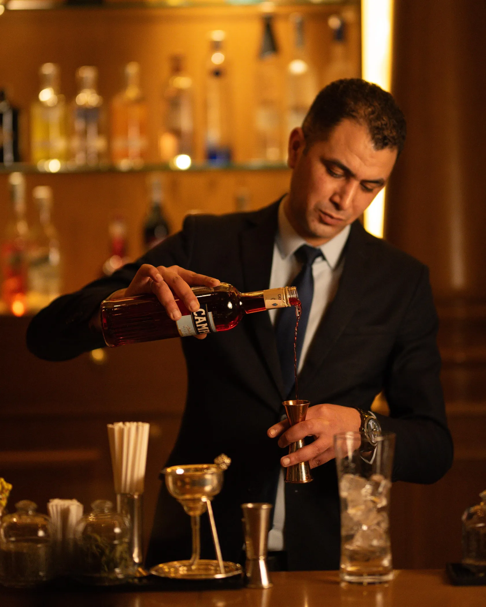 A man in a suit pours a liquid from a bottle into a small metal glass behind a bar.