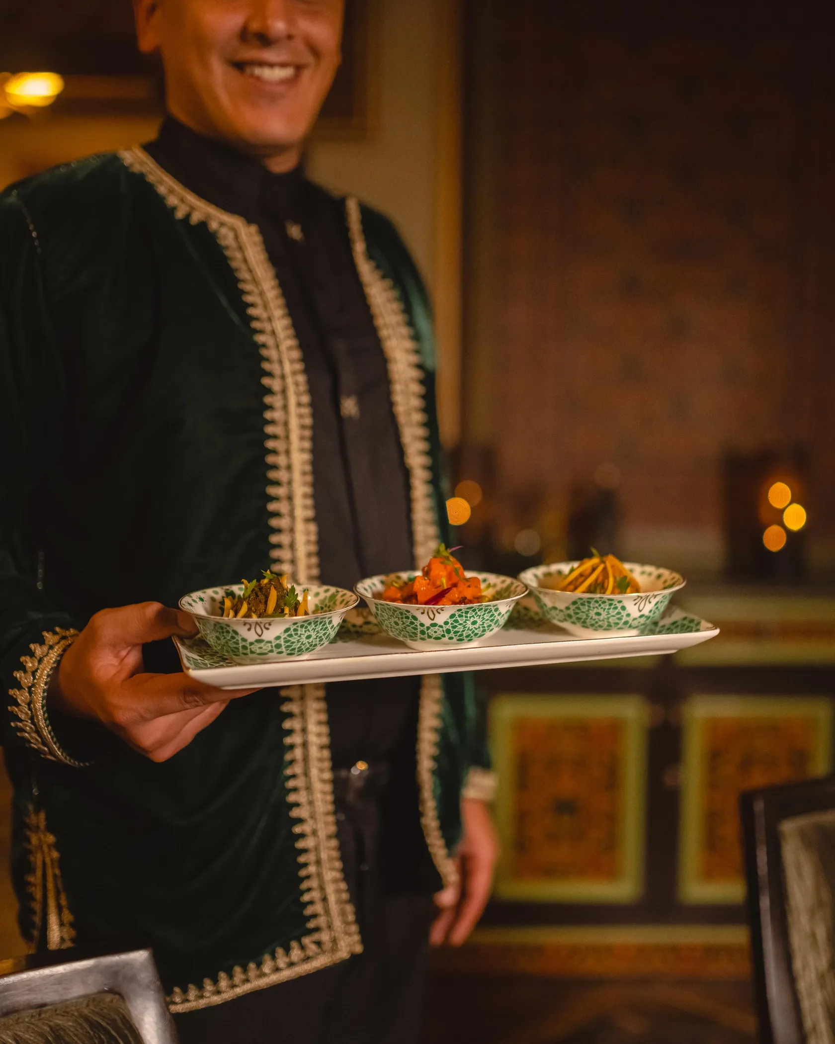 A person in traditional attire carries a tray with three bowls containing vegetables and garnishes.