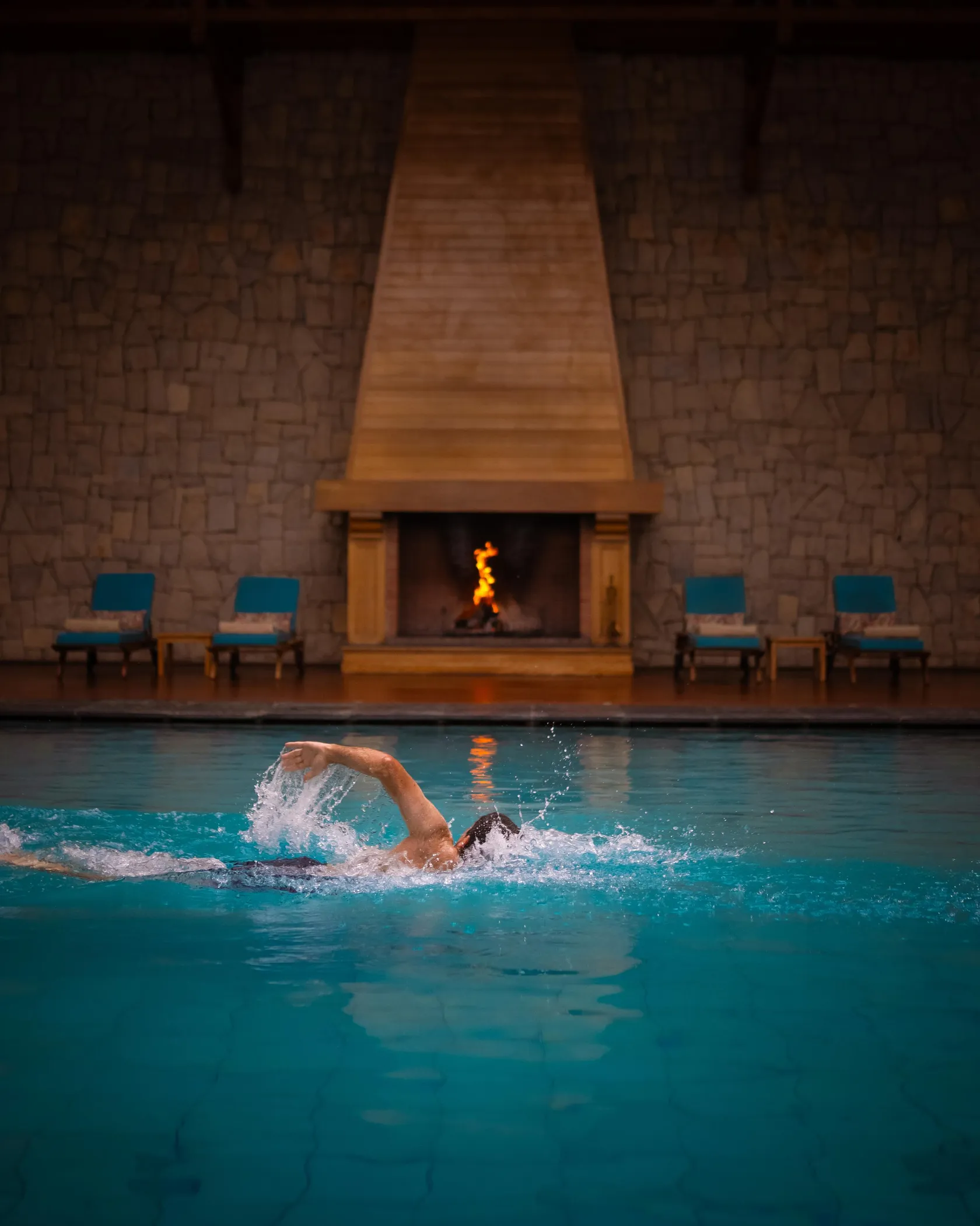 A person is swimming in an indoor pool in front of a lit fireplace.